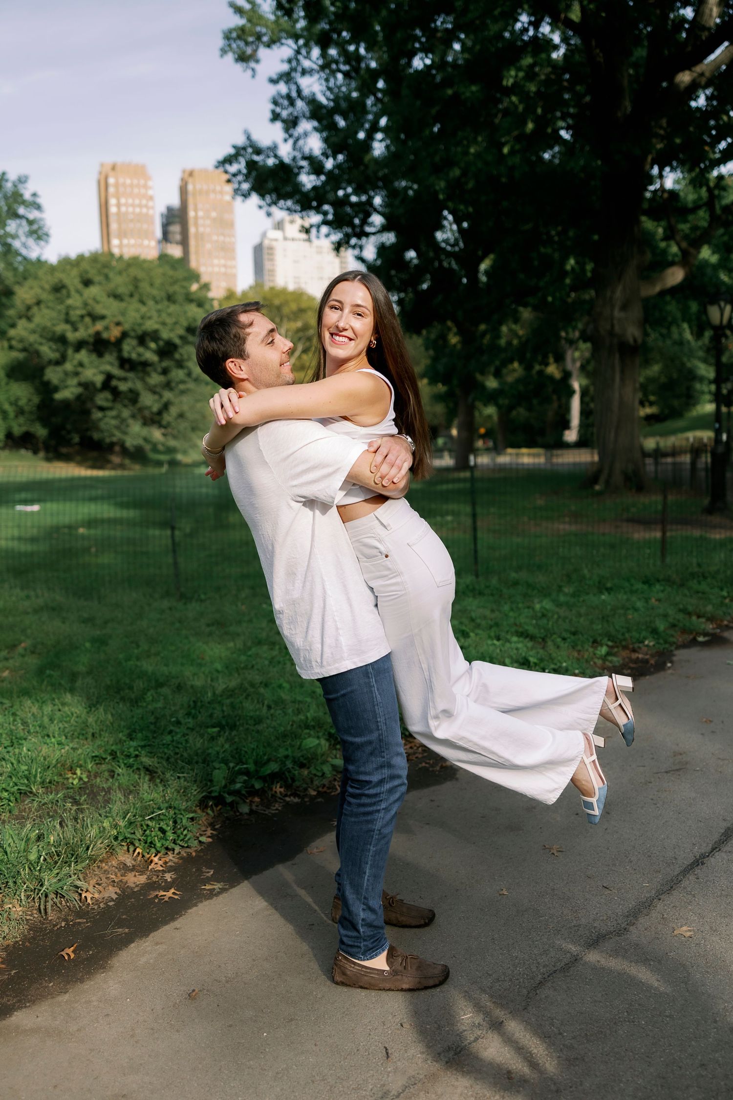 A couple shares a joyful embrace in Central Park with tall city buildings visible in the background.