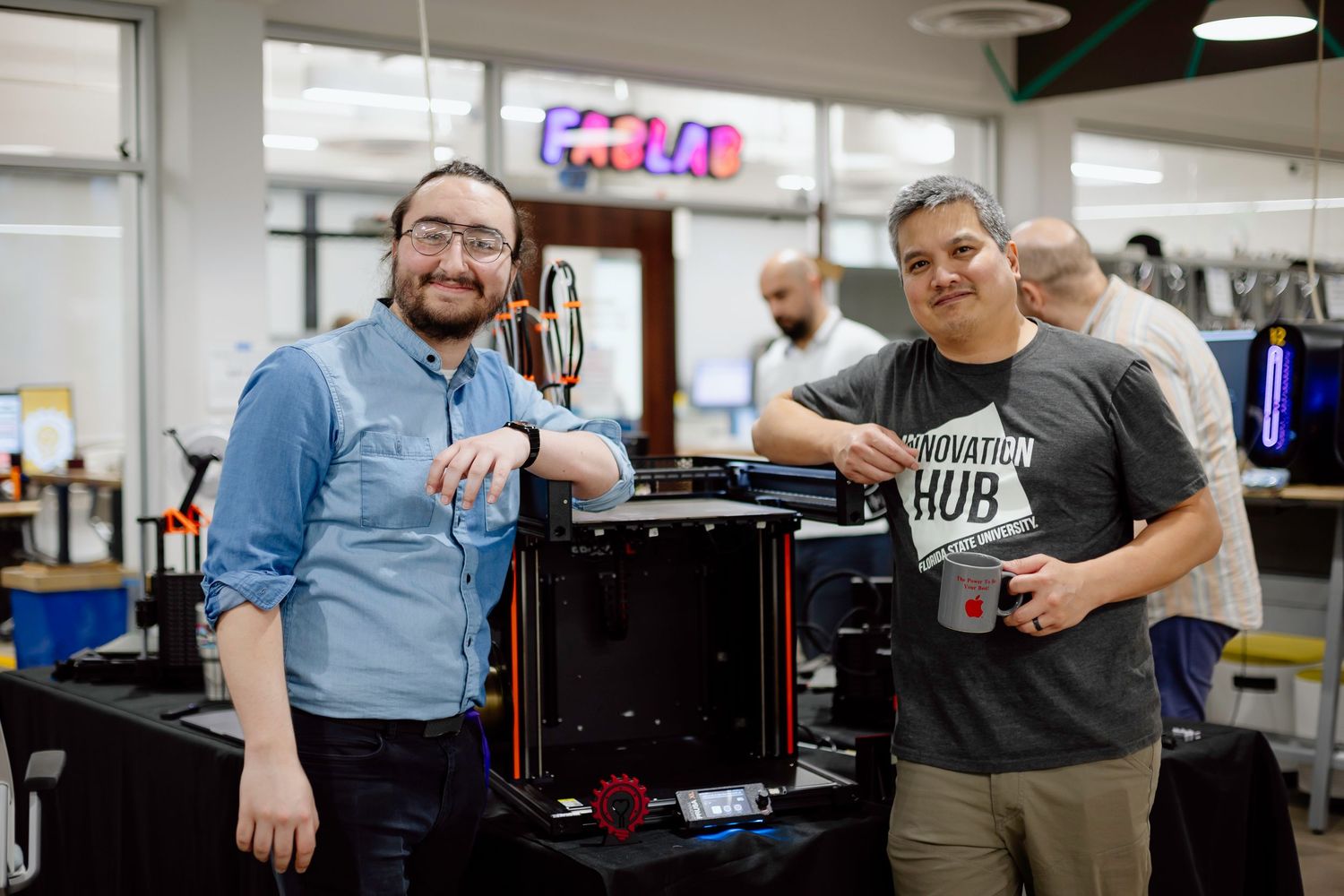Two people are smiling while working together in a research lab or tech workspace next to computer equipment.