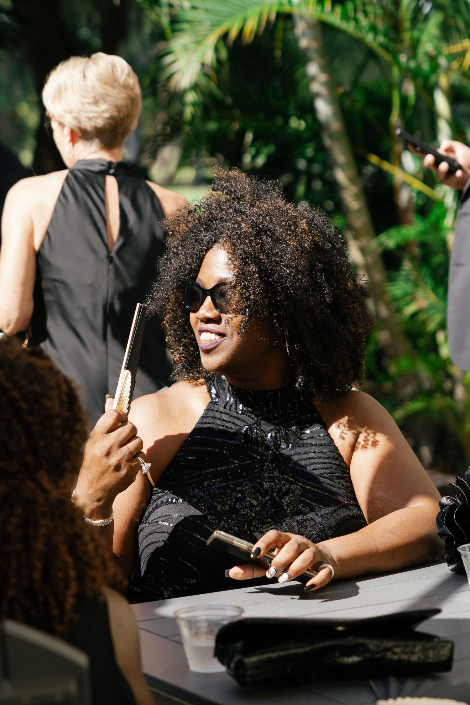 Smiling woman in black dress with sunglasses at outdoor wedding reception table.