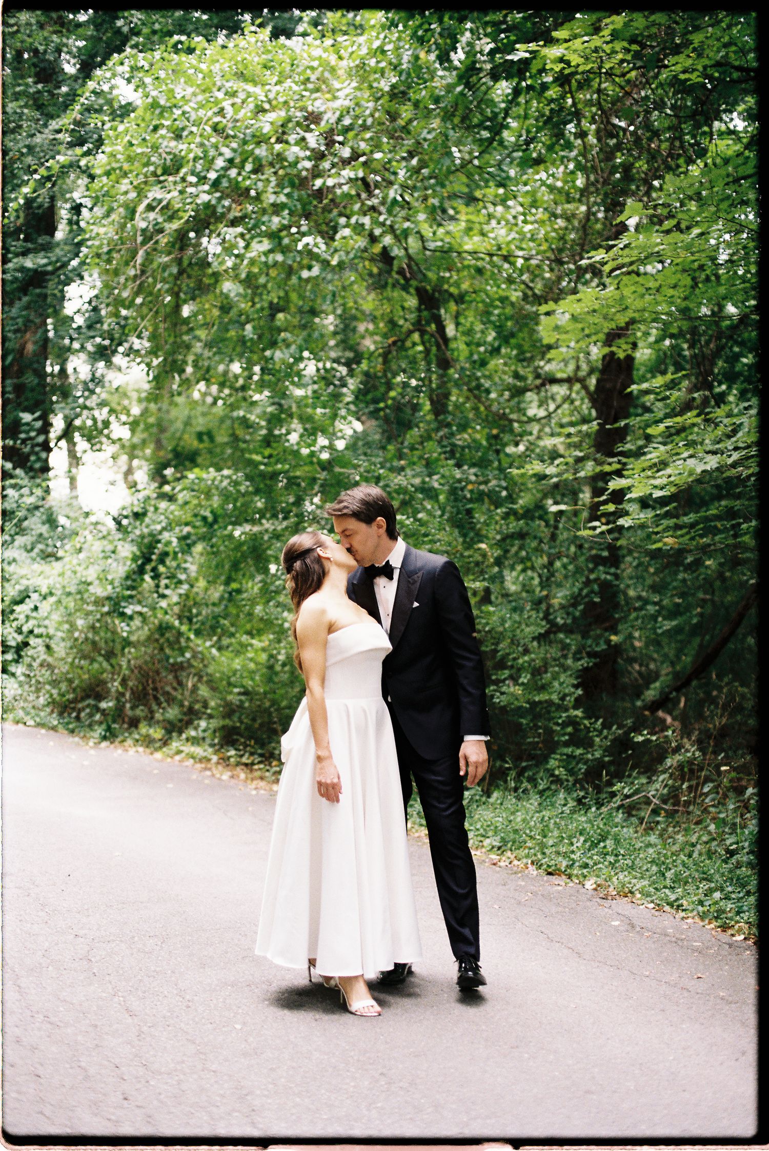 Bride and groom share tender kiss on tree-lined path in lush green setting.