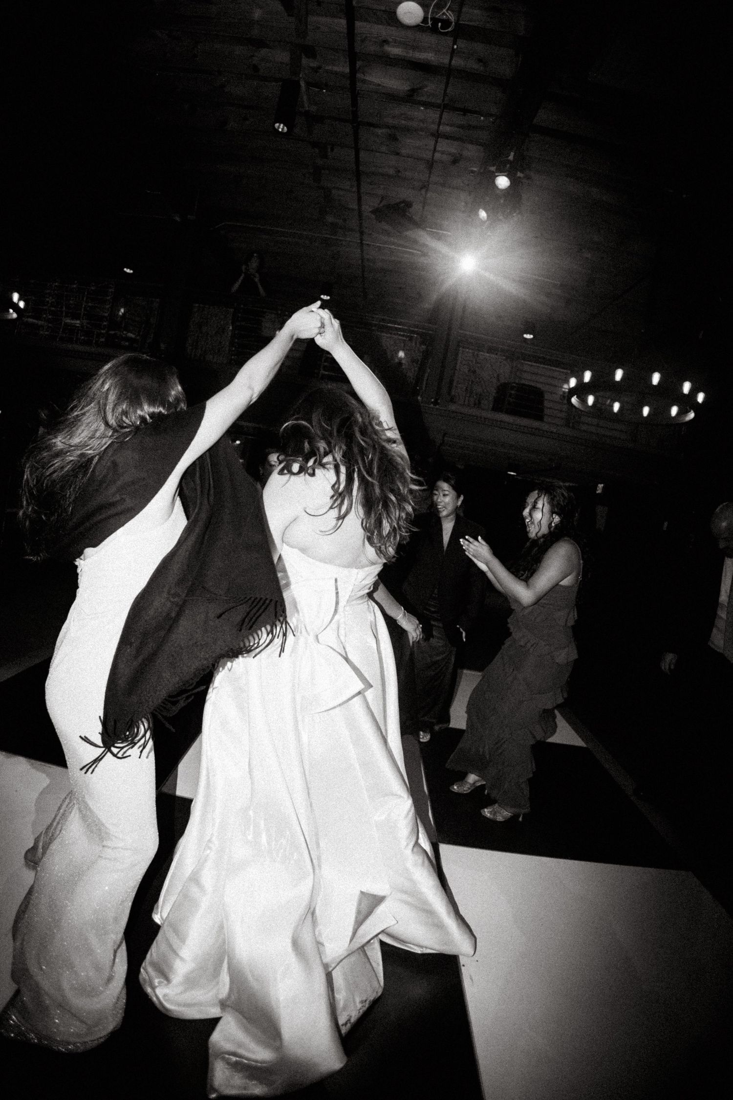 Bride in white gown dancing energetically with guests at a wedding reception in black and white photo.