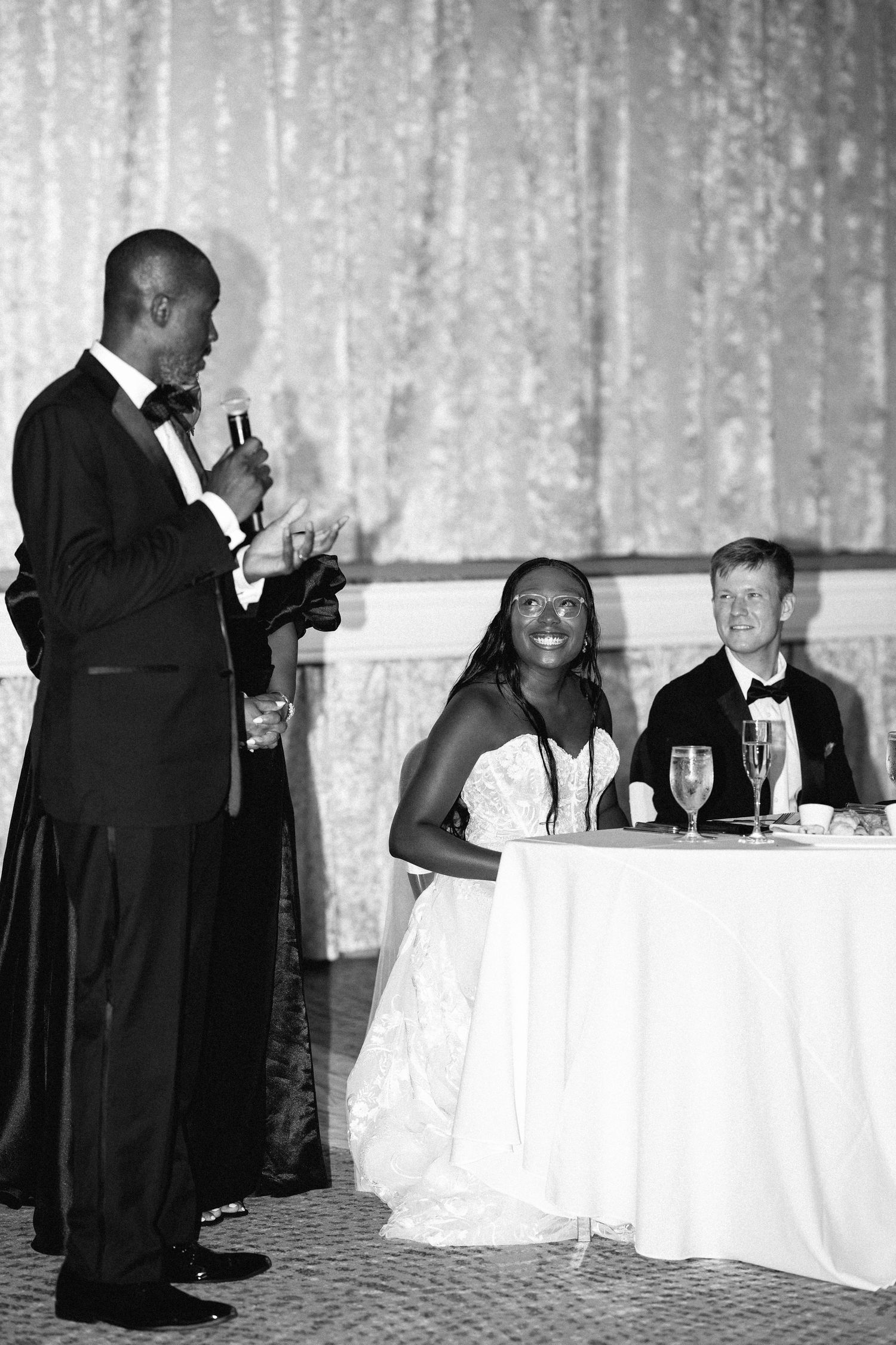 Black and white photo of a man giving a wedding toast as the bride and groom smile at the reception.