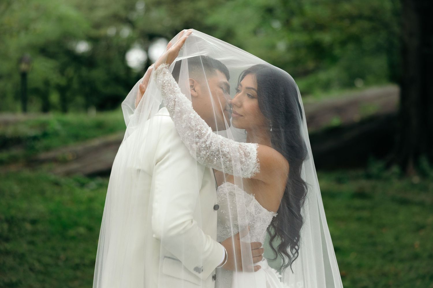 A bride in a white wedding gown and veil poses outdoors in a lush green garden setting.