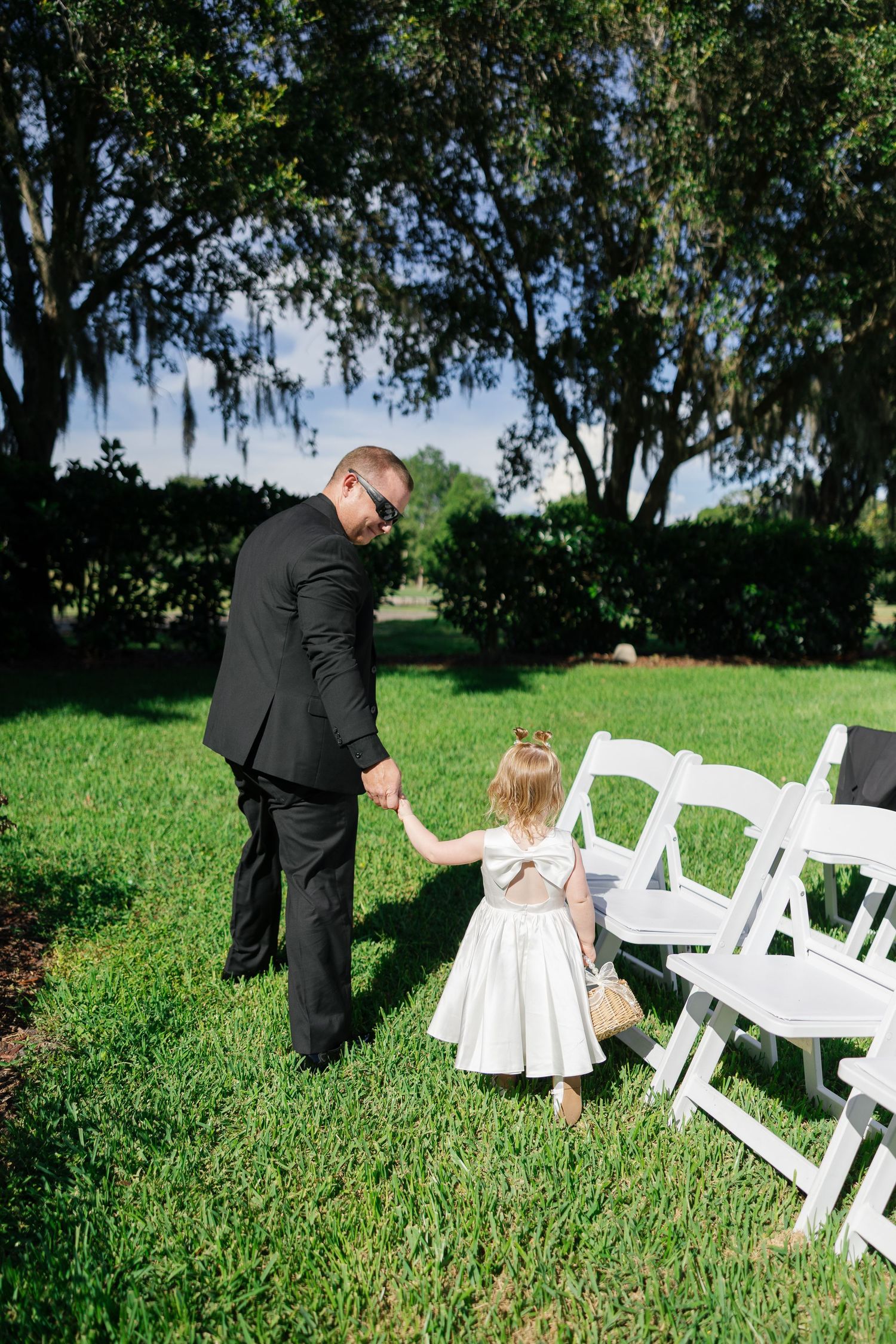 Man in black suit holds hands with young flower girl in white dress at outdoor wedding ceremony.