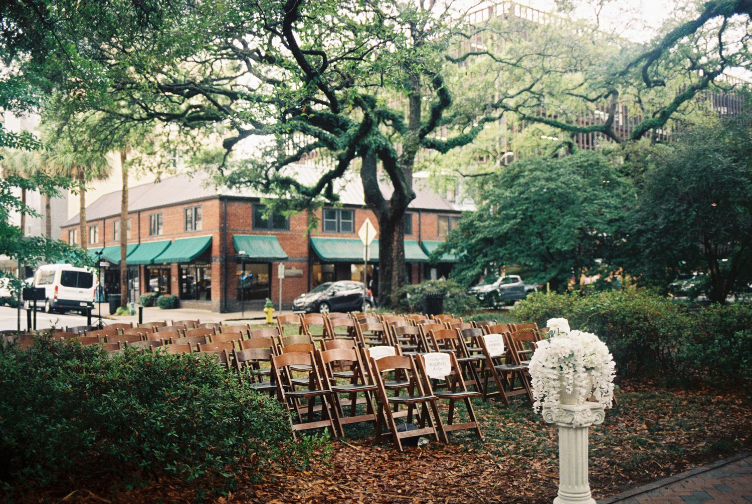 Wooden folding chairs arranged outdoors under large oak trees near a brick building with green awnings for an event.
