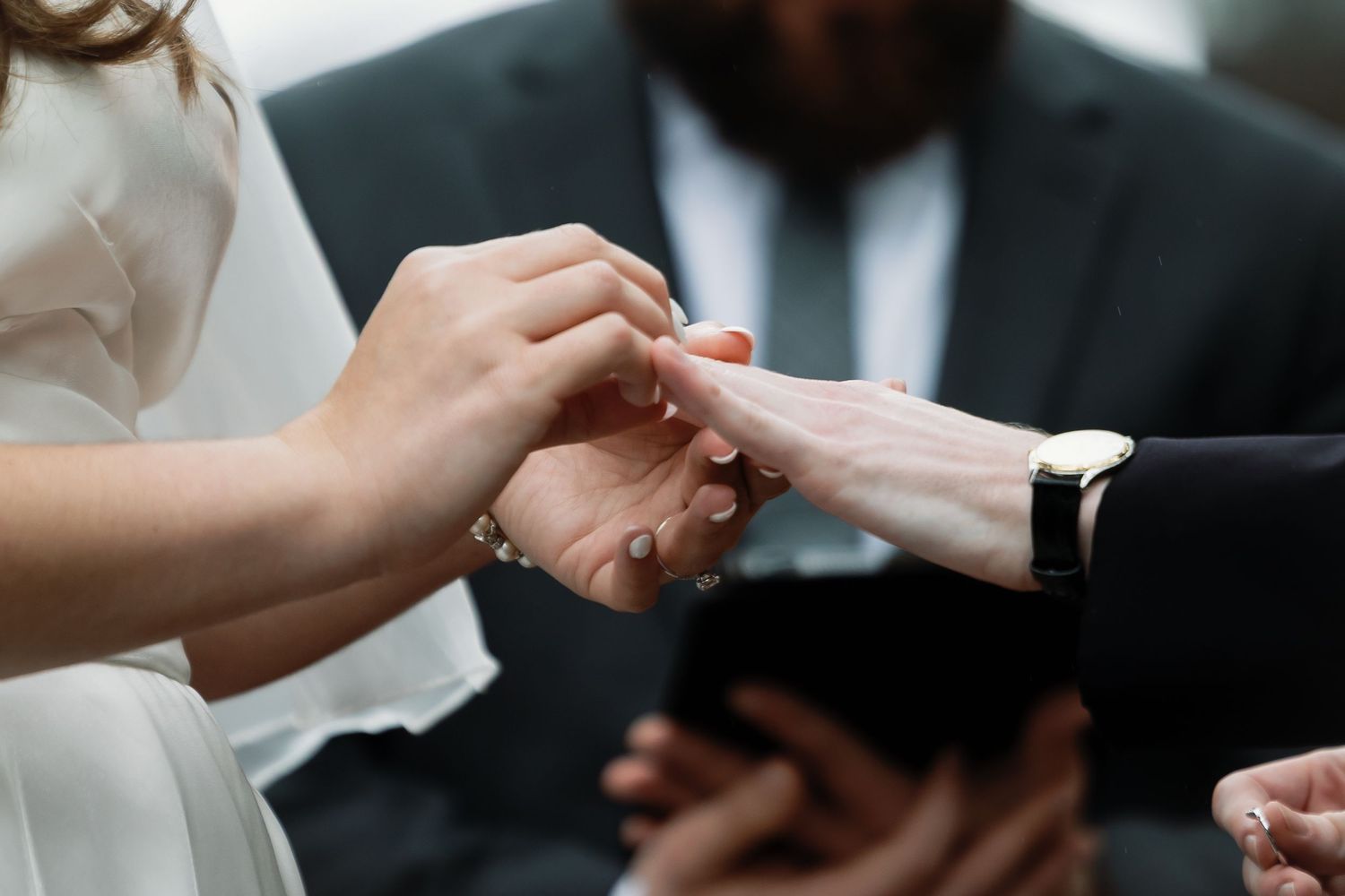 Bride and groom exchanging wedding rings during a ceremony, with an officiant holding a book in the background.