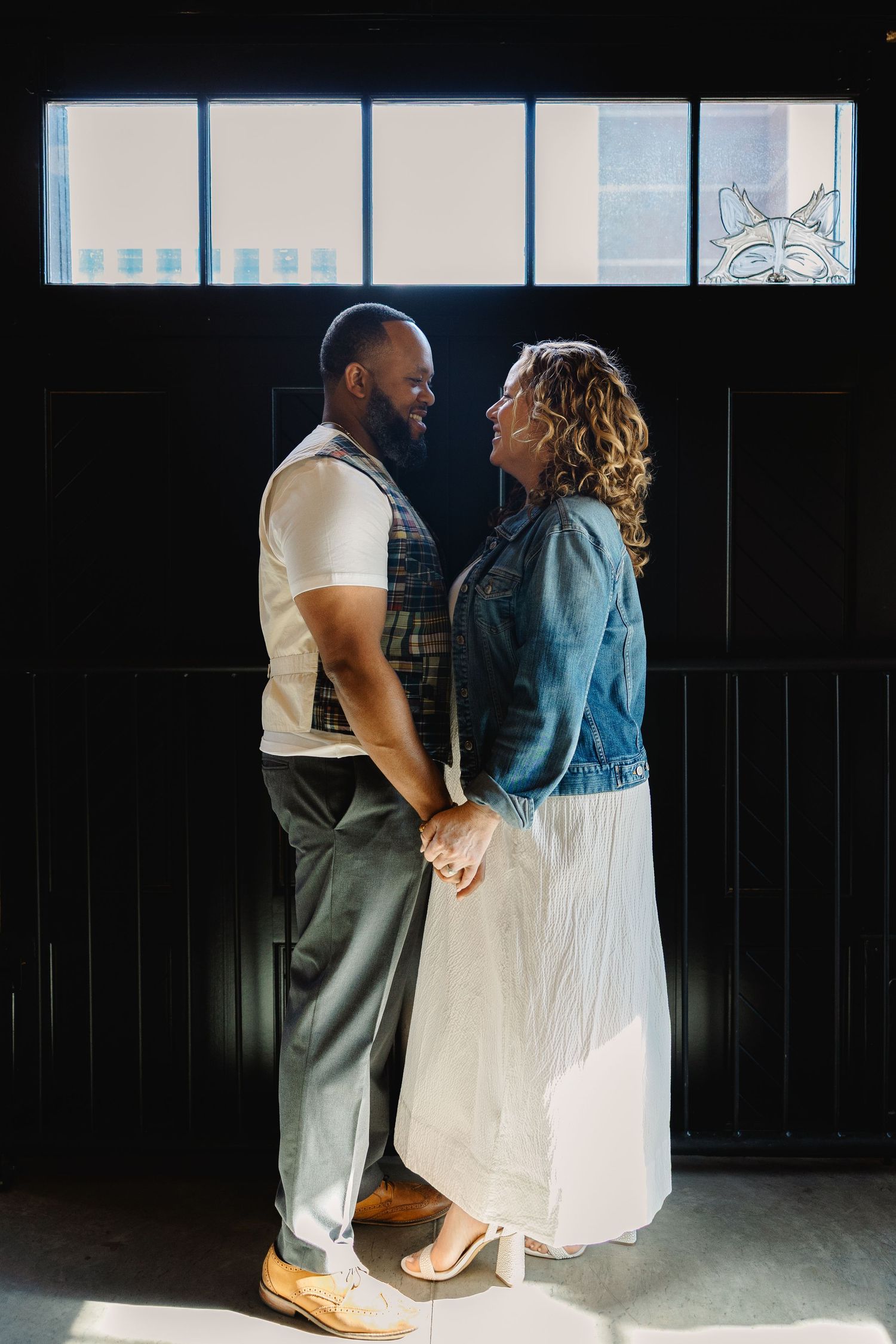 A couple shares an intimate moment in a sunlit room, with the person in a denim jacket and white skirt holding hands with a partner.