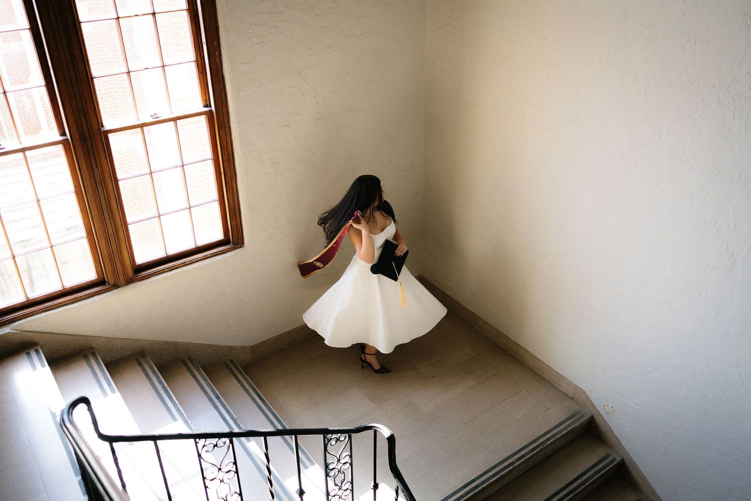 Bride in white dress ascending elegant staircase with wrought iron railing near large window.