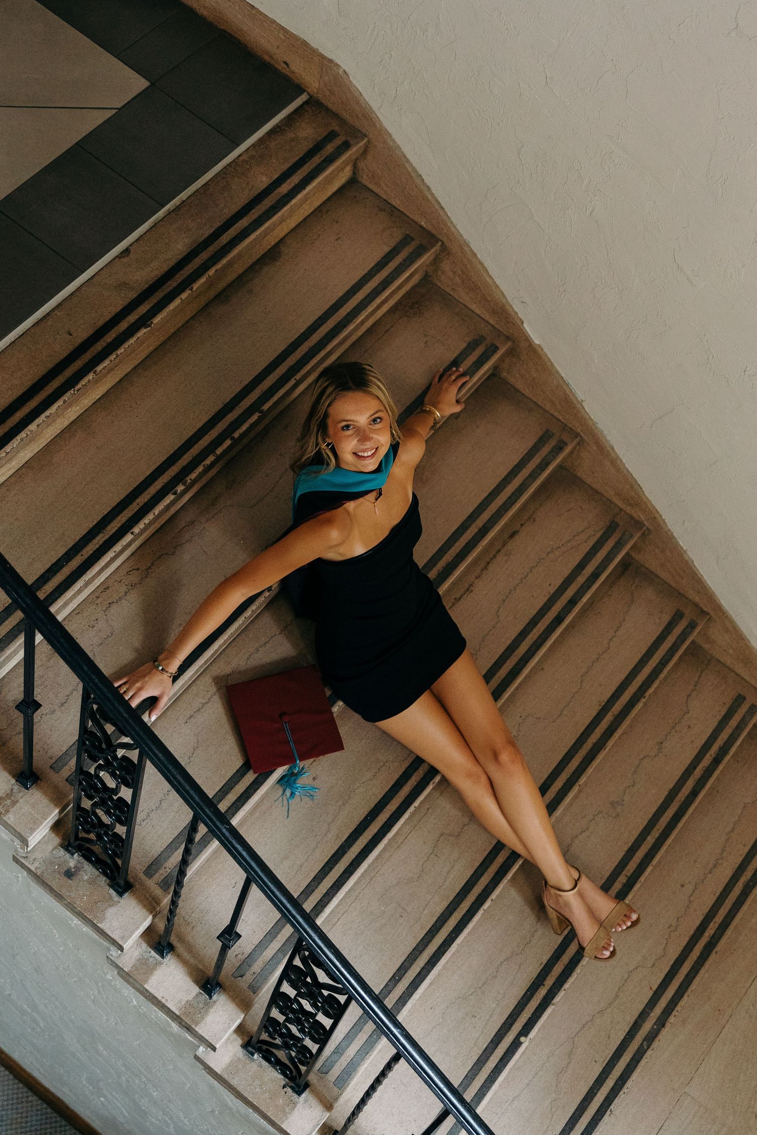 Dancer in black dress and blue scarf strikes dynamic pose on elegant staircase with ornate railing.