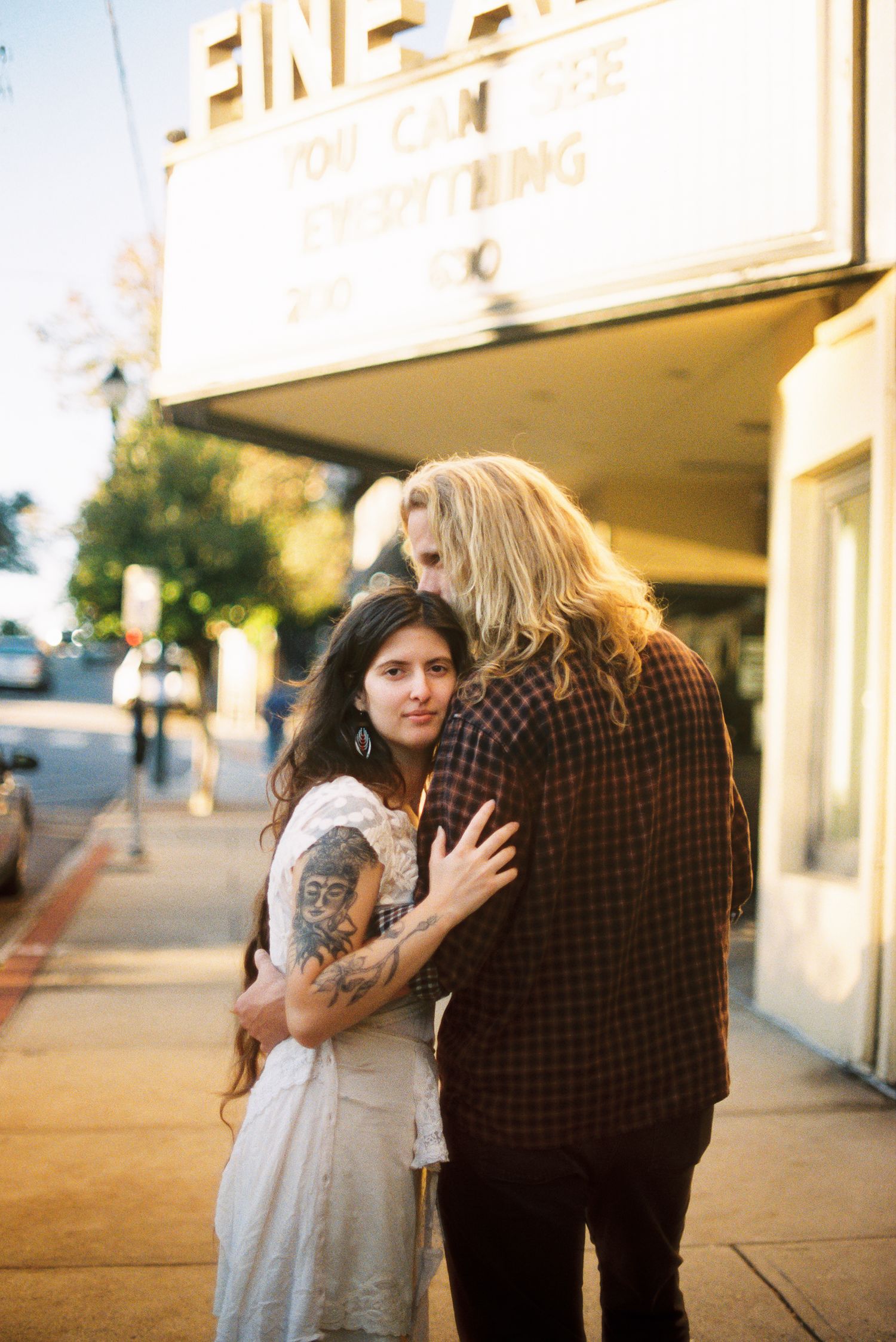 Couple embracing on a sunlit sidewalk beneath a vintage theater marquee, the woman with a tattooed arm glancing over her shoulder.