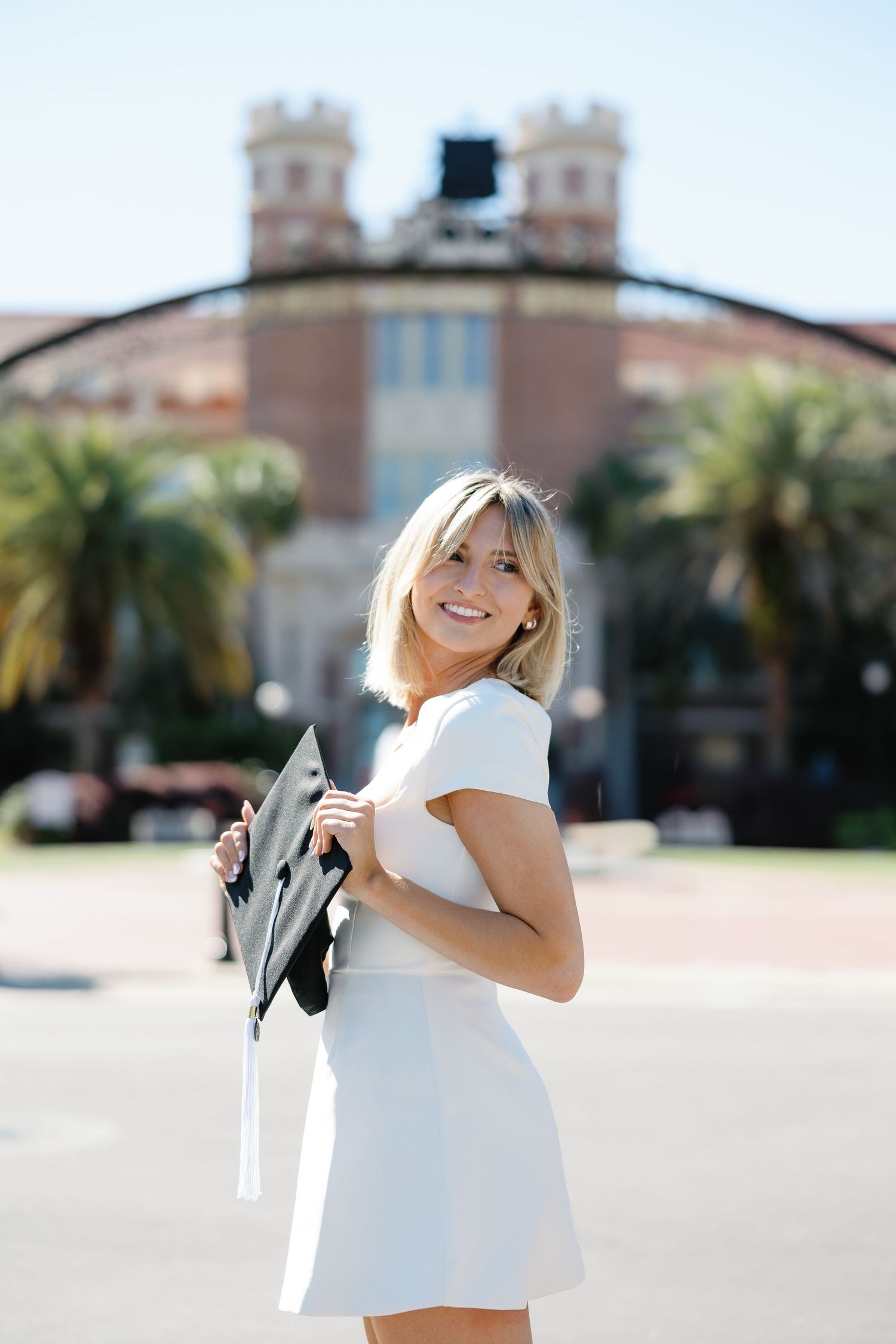 Smiling blonde woman in white dress holding graduation cap outside university building on sunny day.