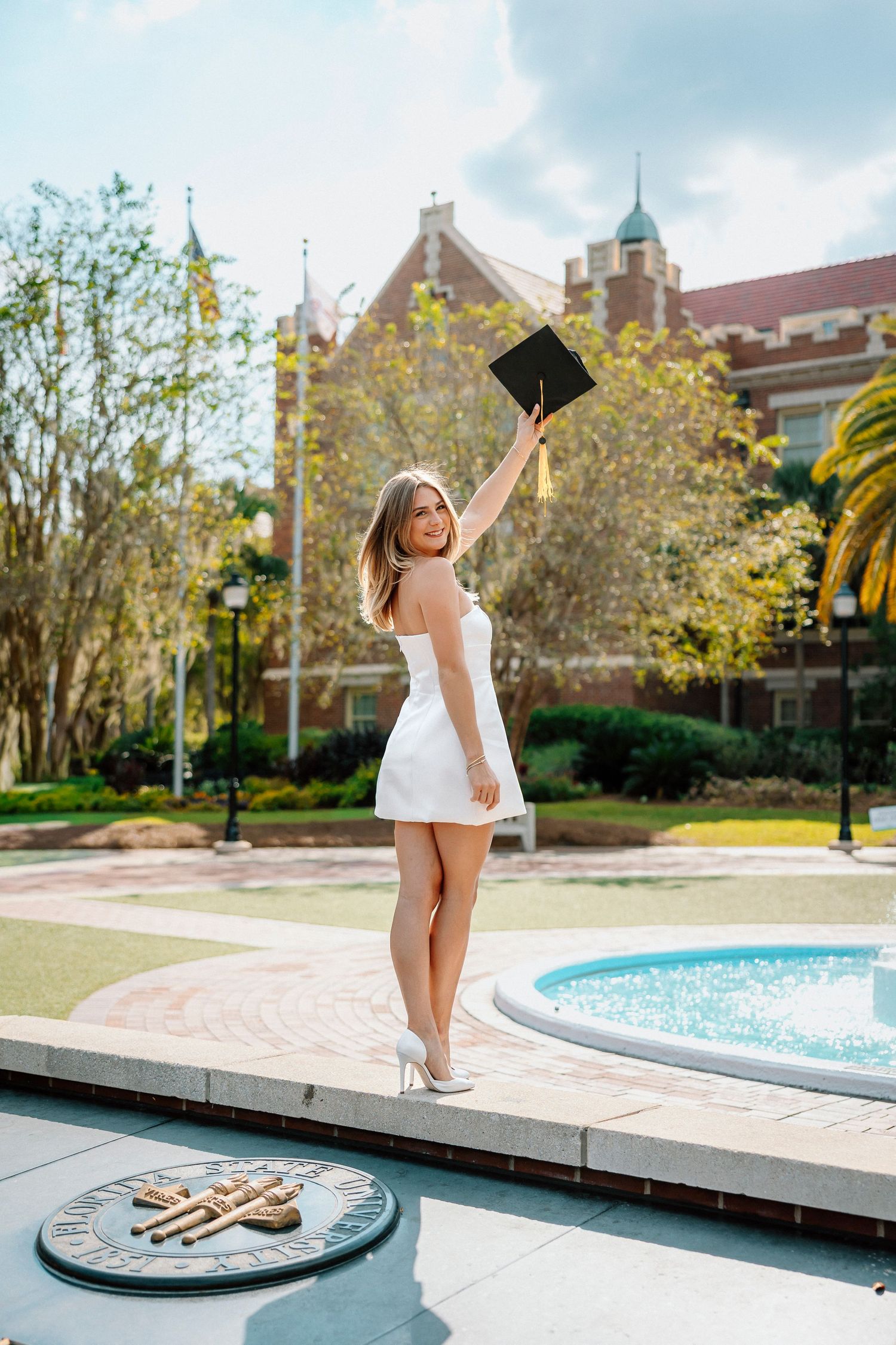Graduate in white dress celebrates on campus near fountain with historic buildings and trees in background.