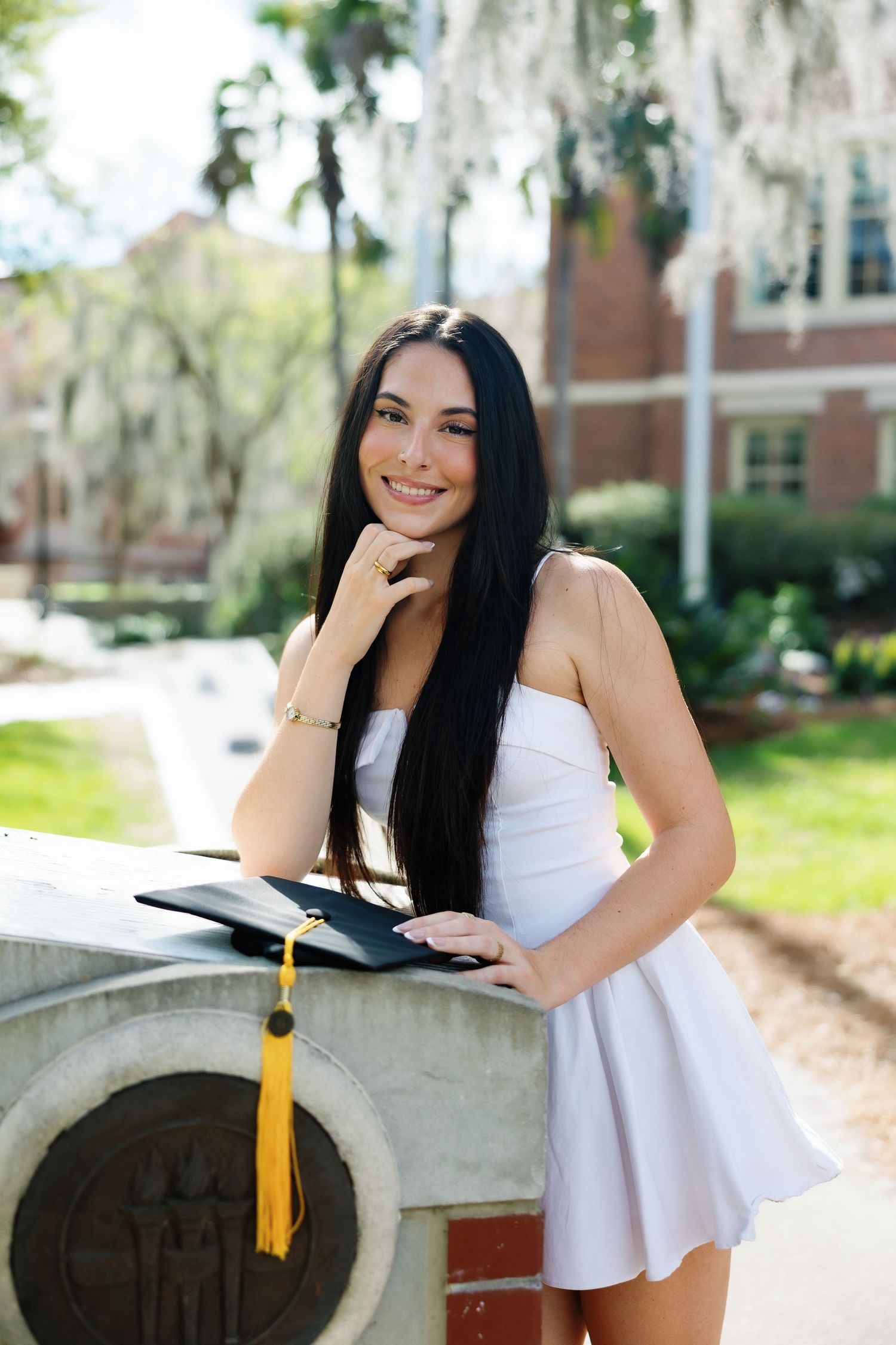Young woman in white dress holding graduation cap and diploma, smiling outdoors on college campus.