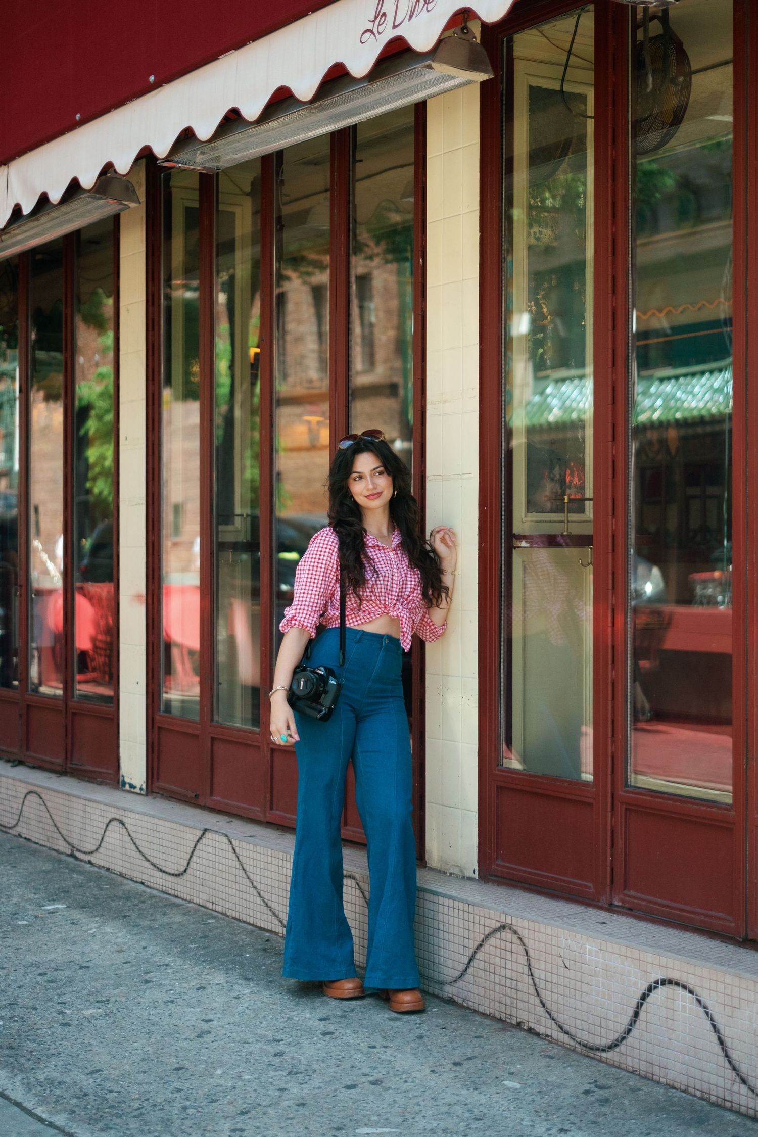 Woman in pink top and blue flare pants posing outside a red-framed restaurant storefront on a city sidewalk.