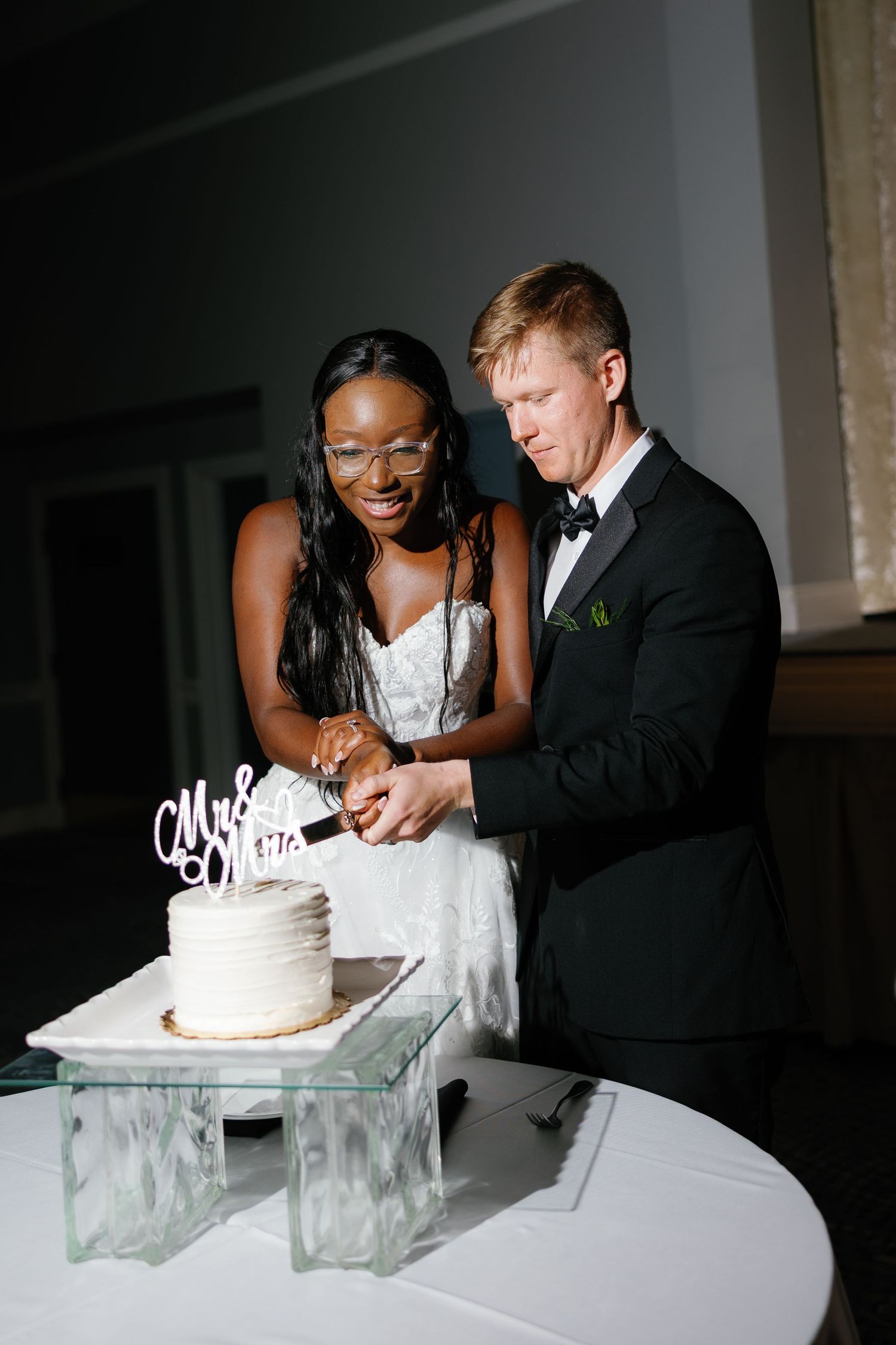 Interracial couple cutting white wedding cake together, bride in lace dress with glasses, groom in black tuxedo.