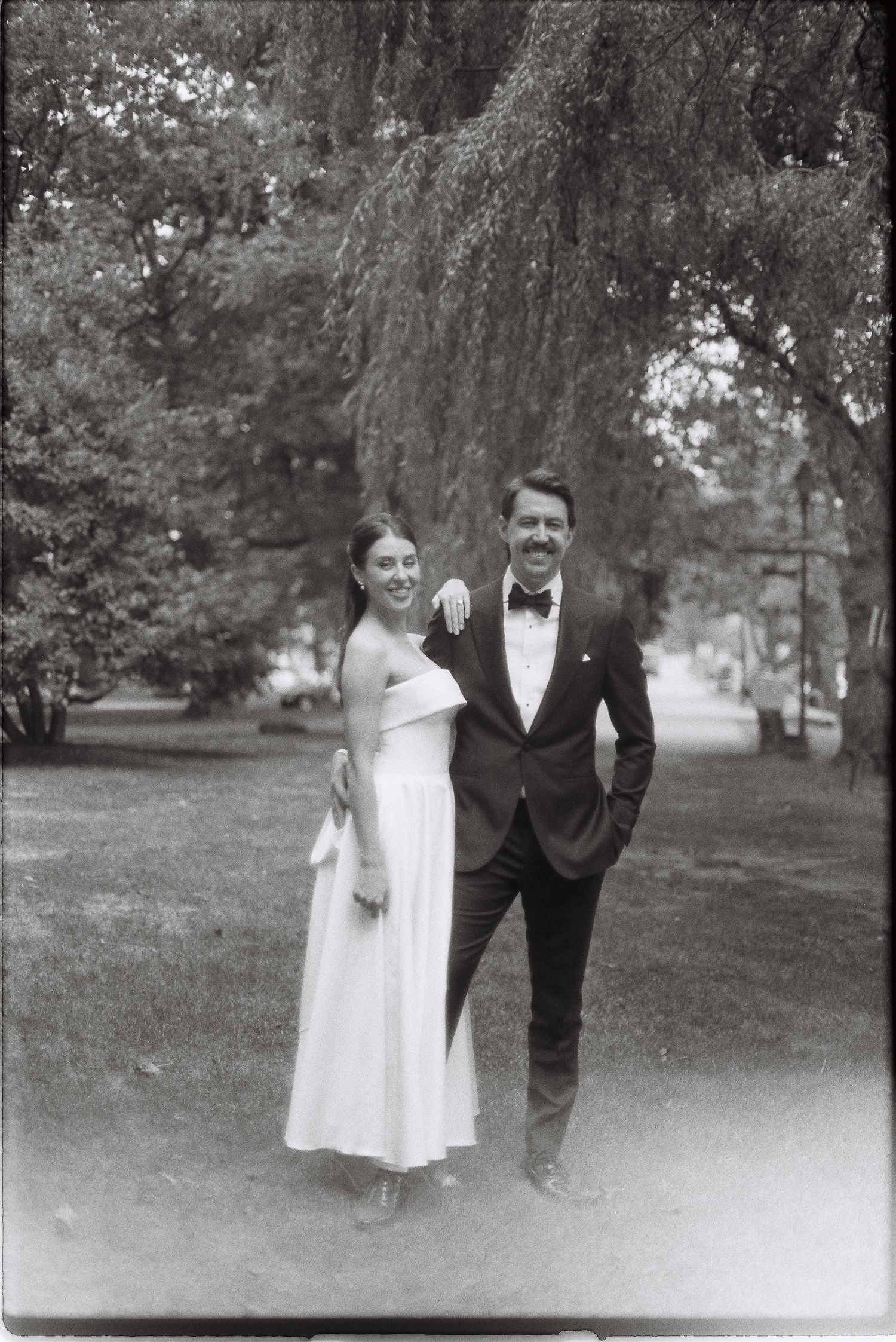 Bride and groom pose together under a weeping willow tree in black and white wedding portrait outdoors.