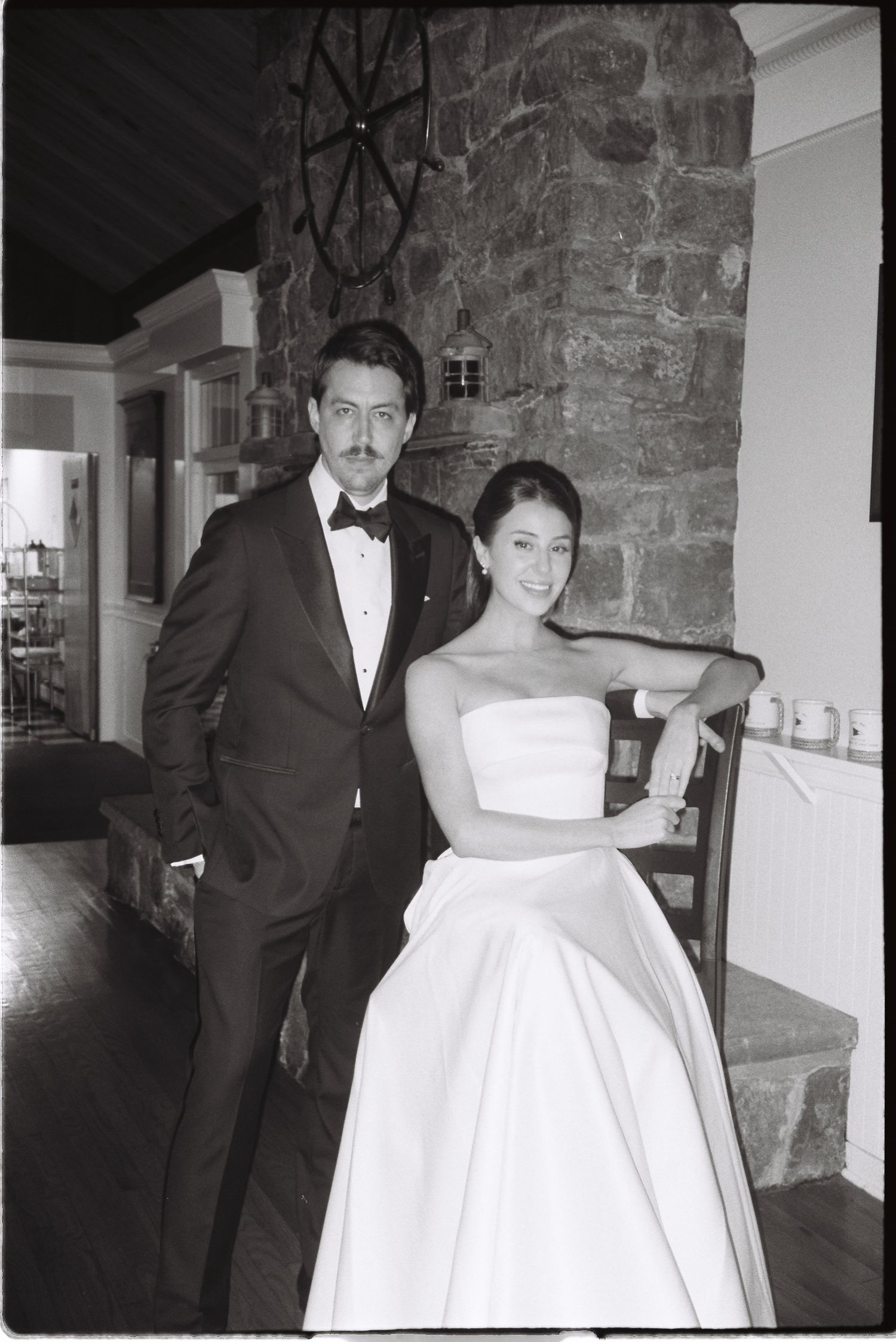 Black and white photo of a couple in wedding attire posing indoors near a stone wall.