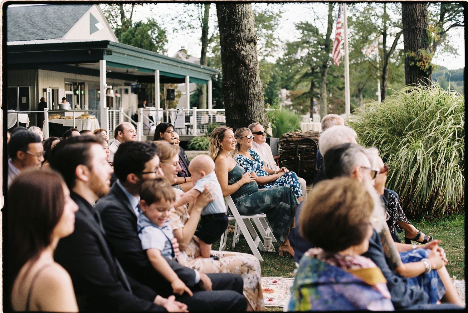 Guests seated outdoors at a wedding ceremony on a sunny day, with a house and American flag visible in the background.