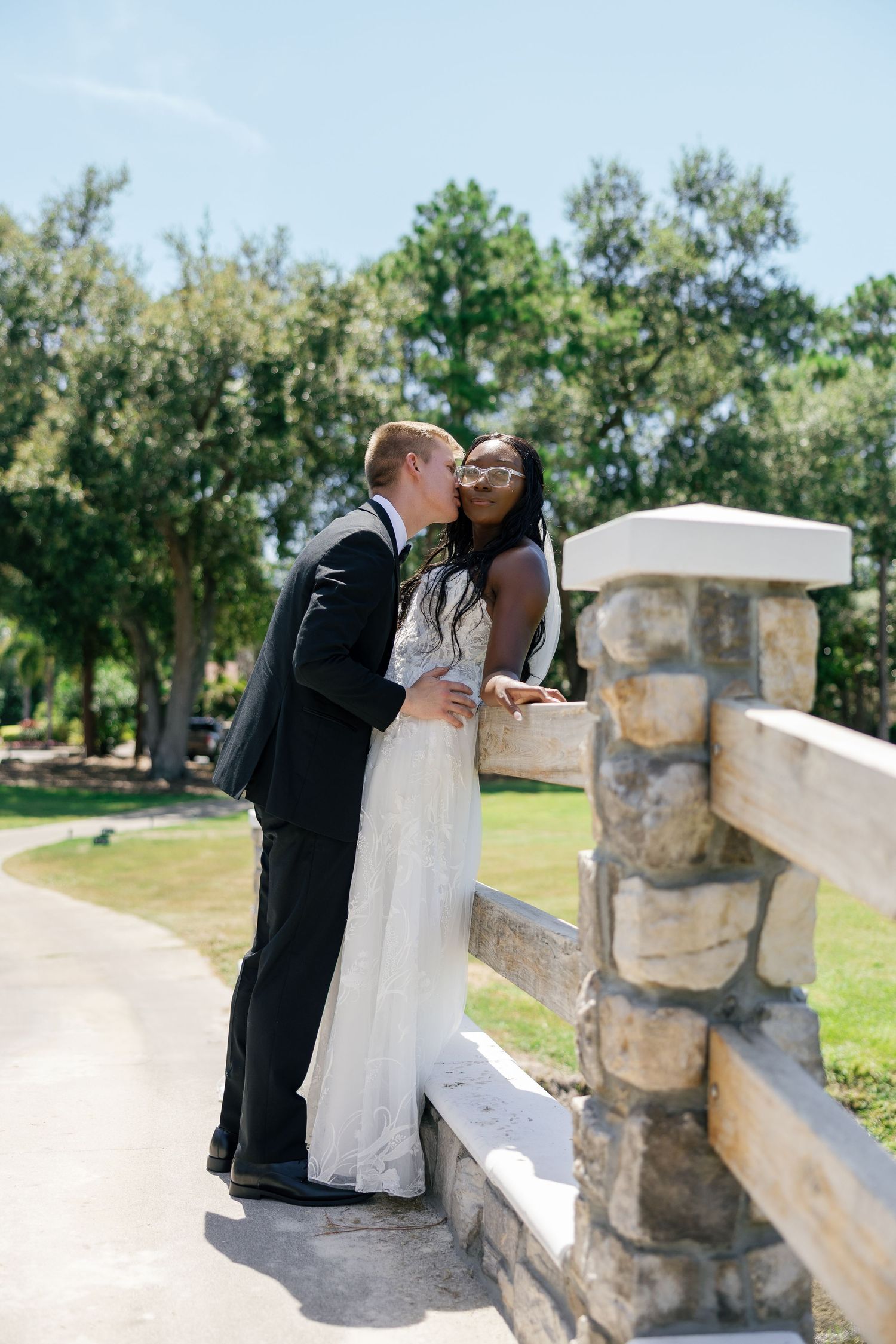 Interracial bride and groom share romantic kiss by stone fence surrounded by lush green trees outdoors.