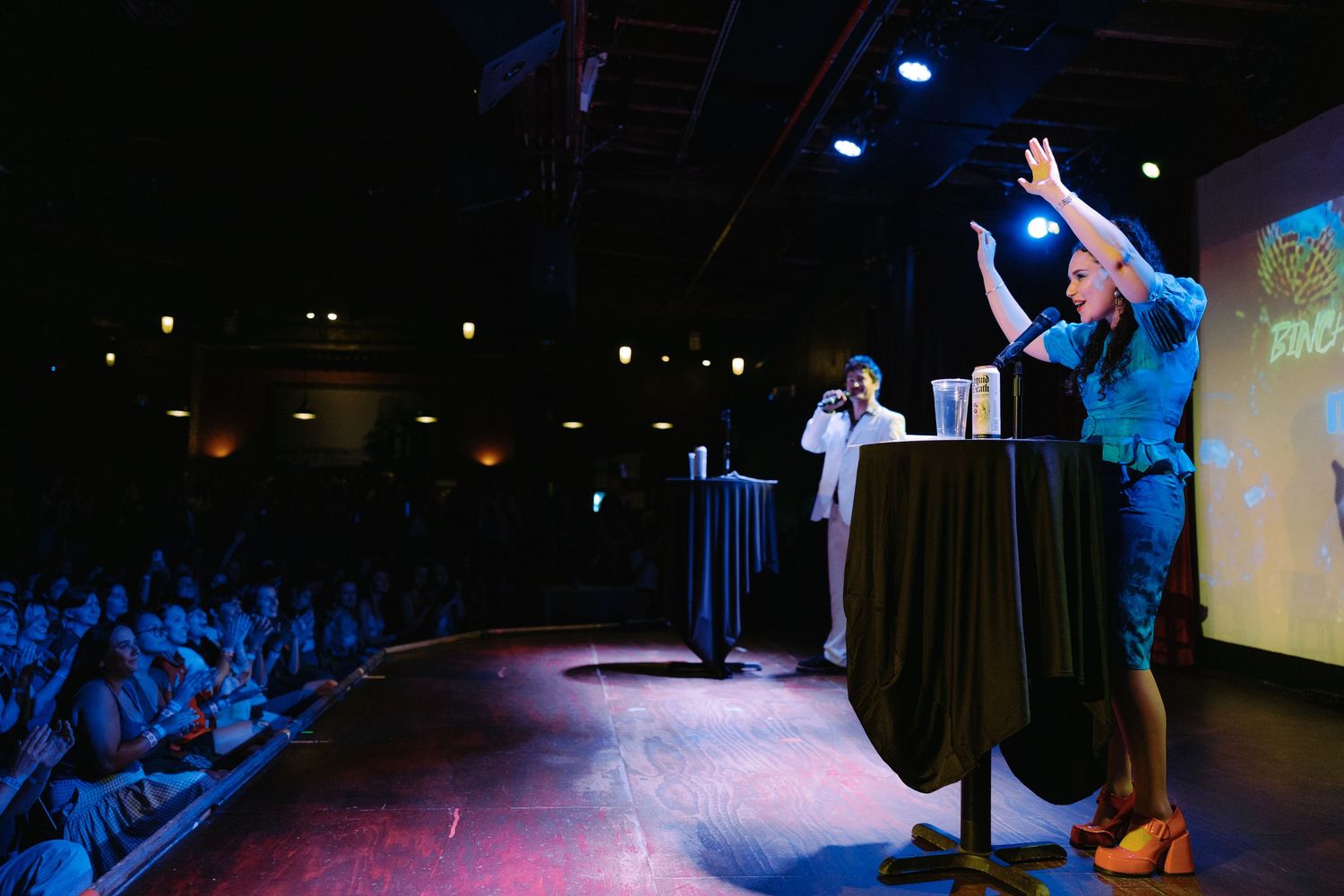 Two speakers present on a dark stage before a large audience, with blue lighting and a projection screen visible.