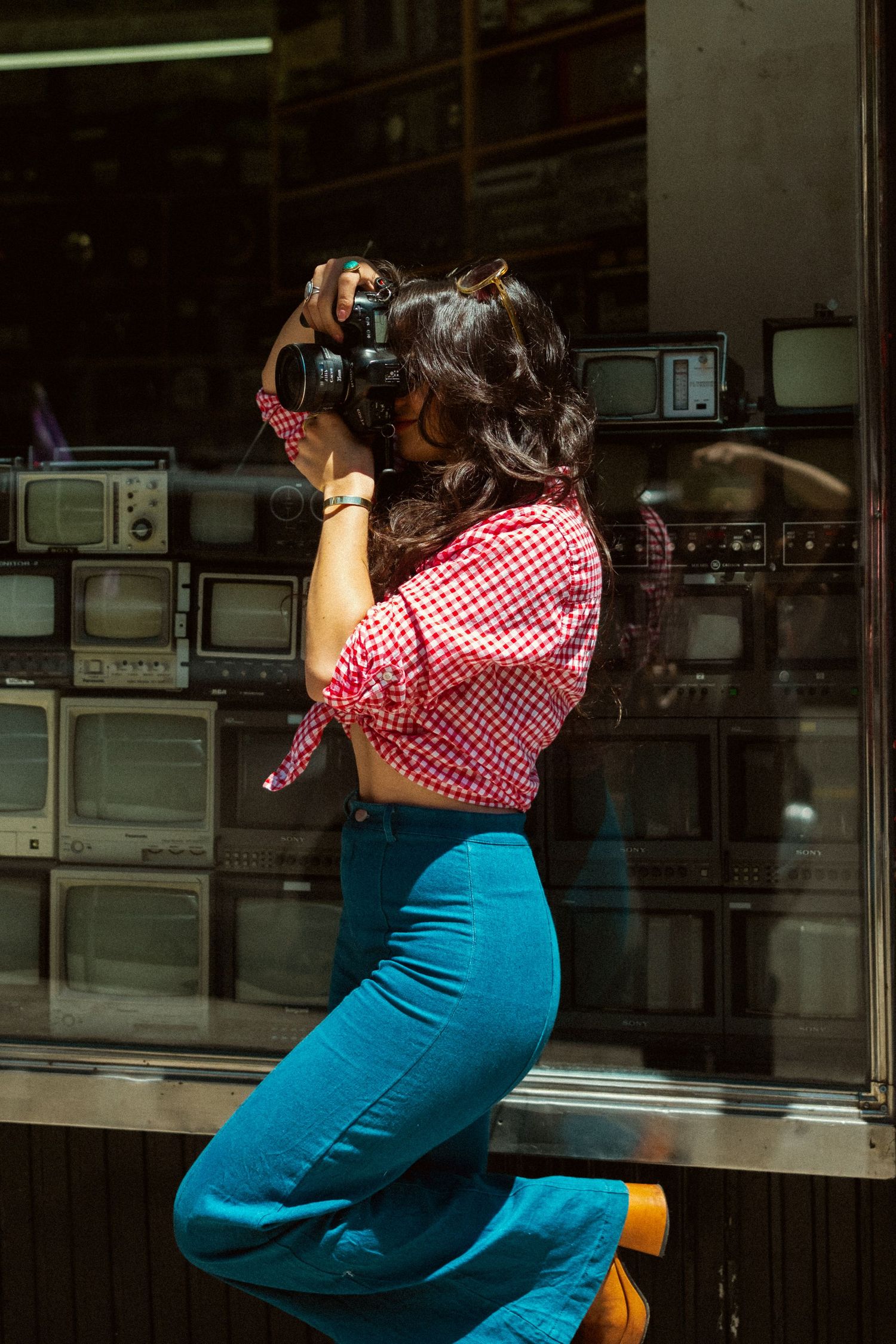 Woman in red checkered top and blue flared pants photographs with DSLR camera in front of vintage TV store window.