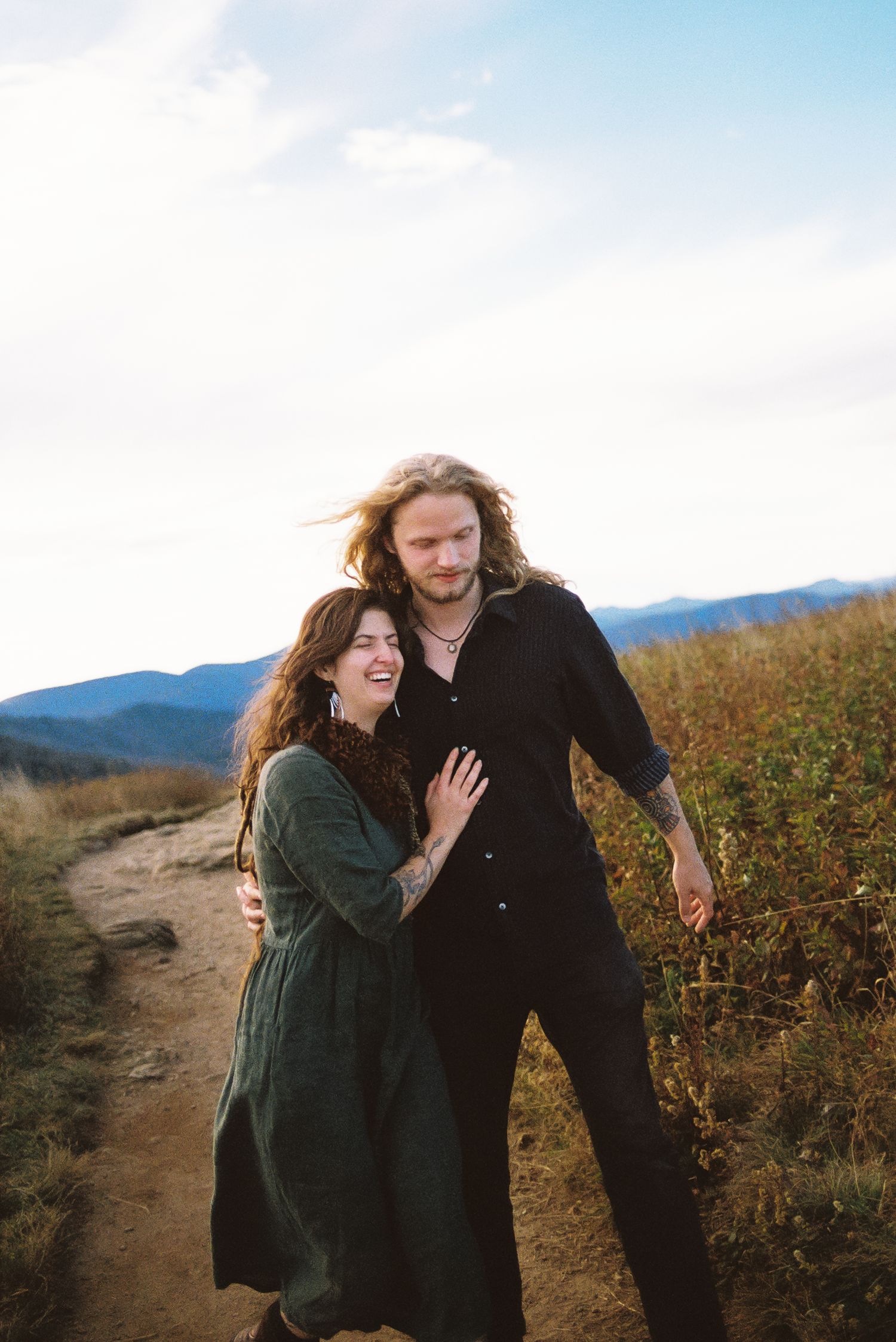 A smiling couple embraces on a mountain trail, with a woman in a green dress leaning on a long-haired man in black.