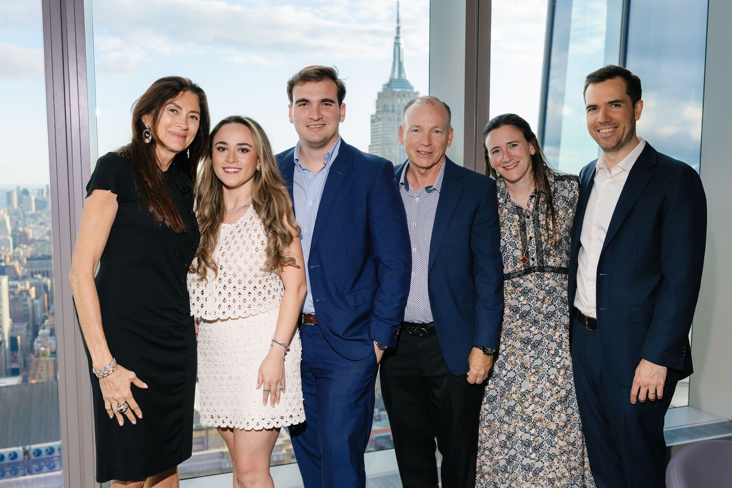 Six professionals pose together smiling at a rooftop event with the Empire State Building visible through windows behind them.