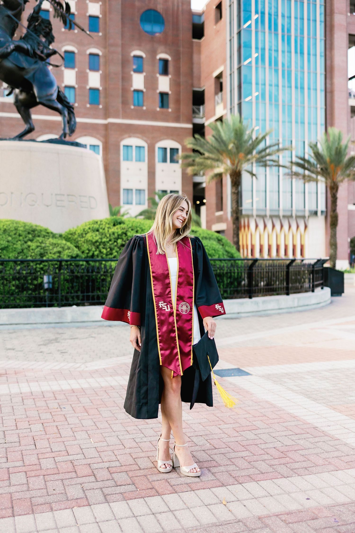 Graduate in cap and gown poses for graduation photos on brick campus courtyard with palm trees.
