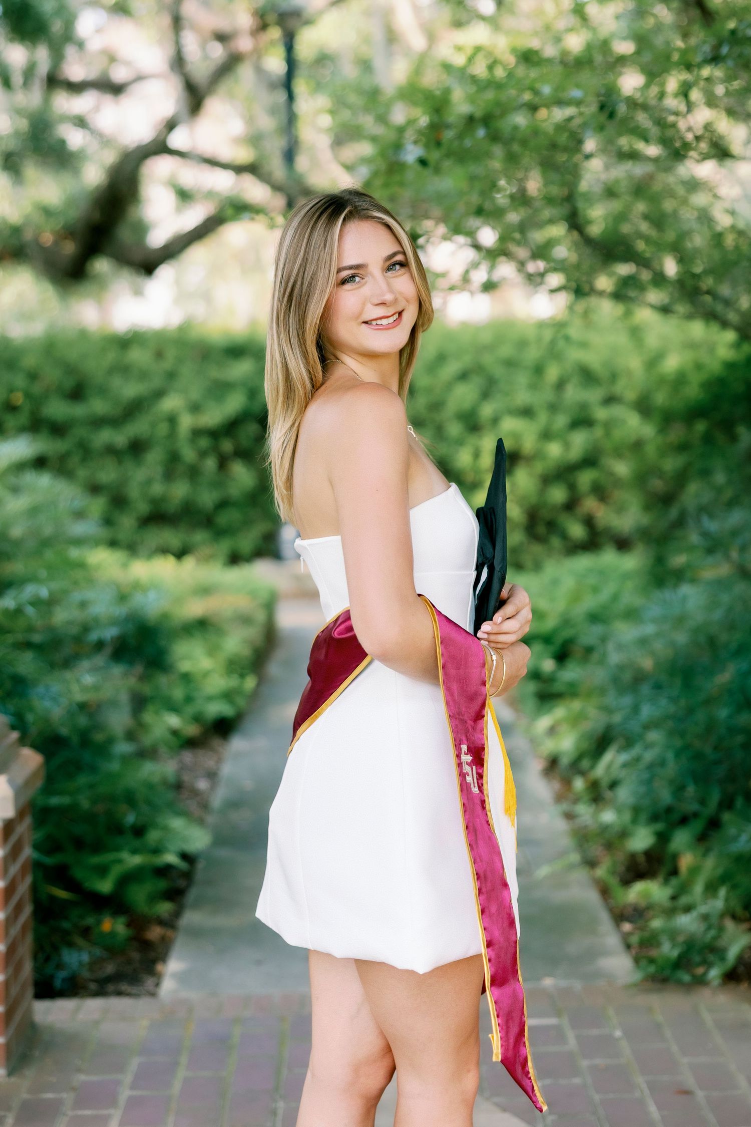 A person in a white dress poses outdoors on a brick path surrounded by lush green foliage in a garden setting.