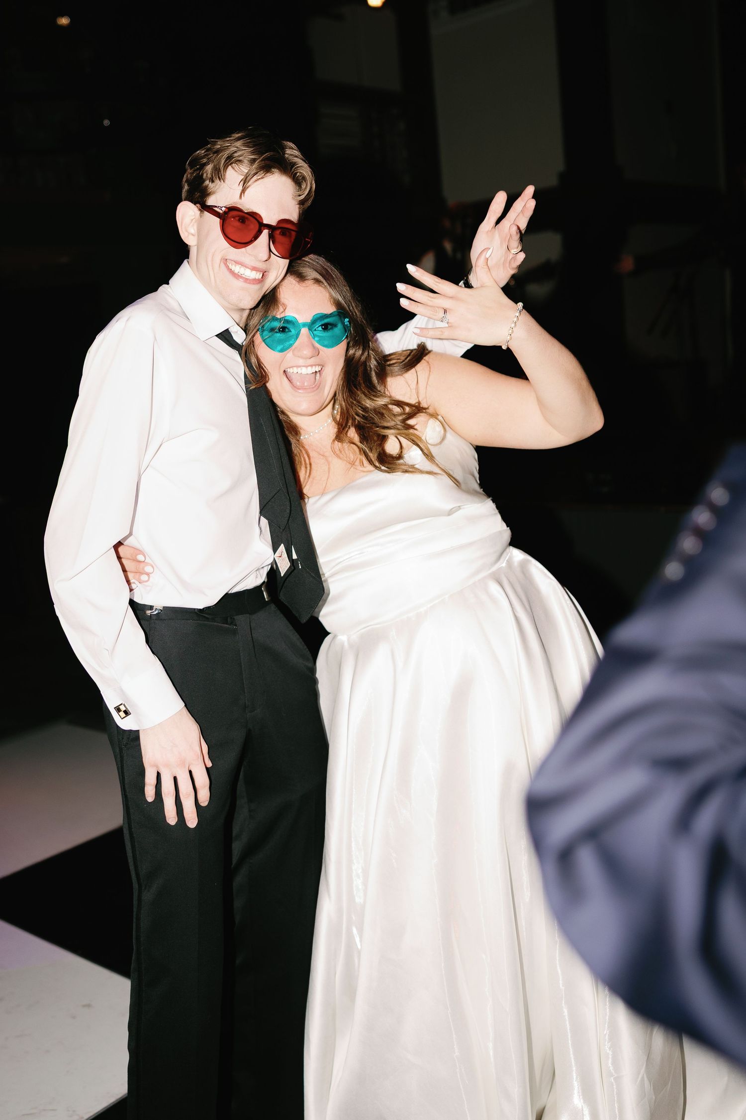 Joyful bride and groom wearing colorful heart-shaped sunglasses dancing and celebrating at their wedding reception.