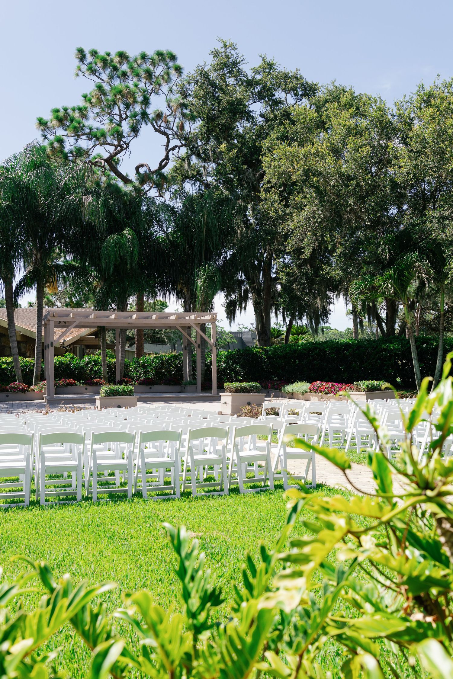 Outdoor wedding ceremony setup with white folding chairs on green lawn beneath lush trees and a wooden pergola altar.