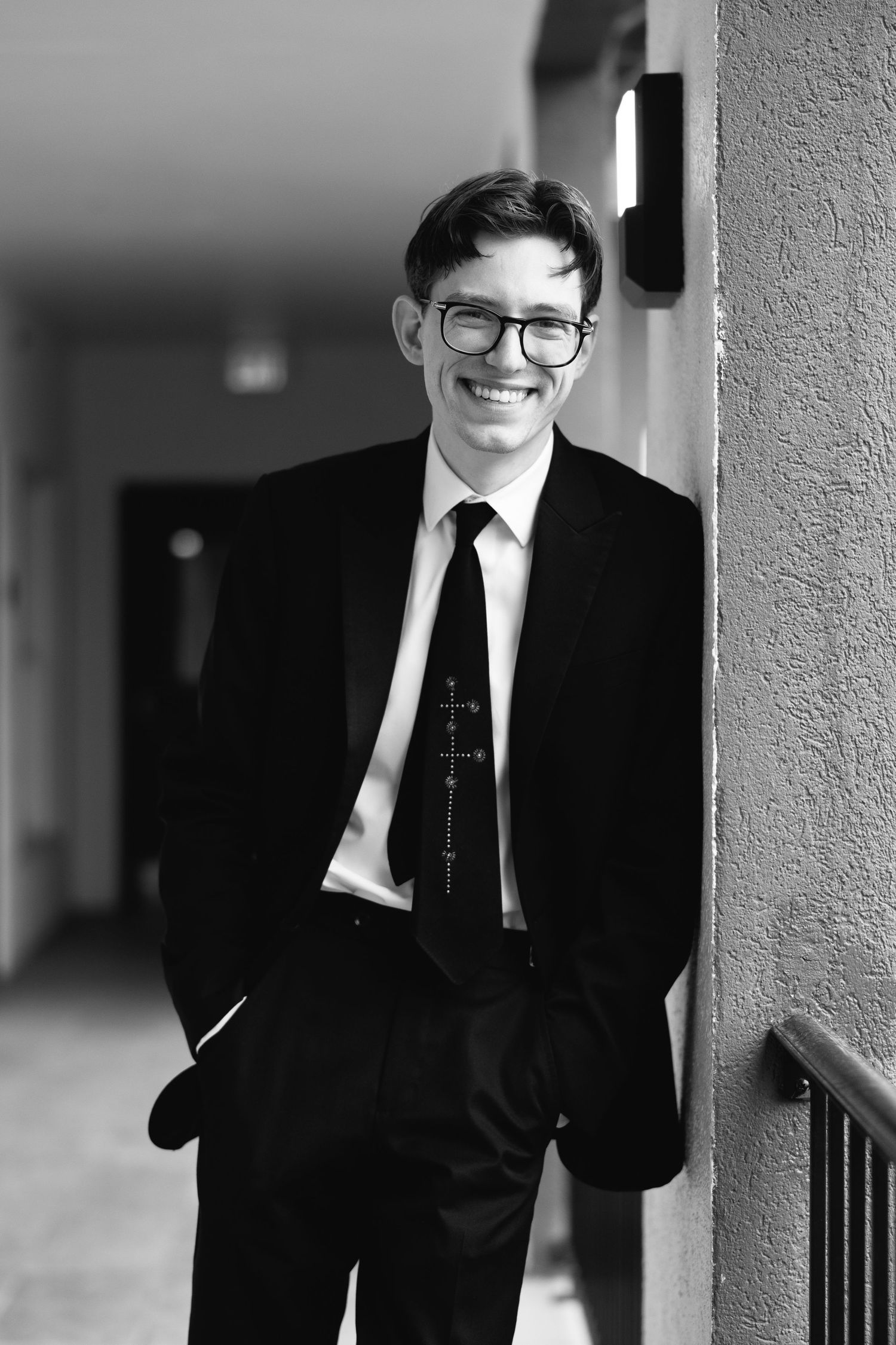 Young man in black suit and tie with glasses smiling confidently, leaning against a wall in a hallway.