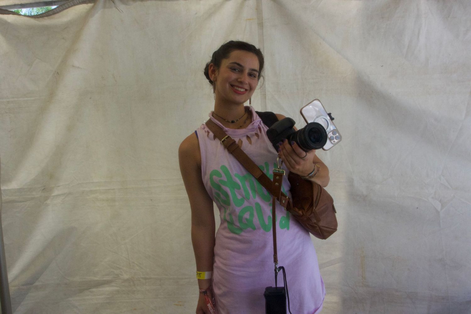 Someone in a light pink dress holds a camera and smiles while standing against a white backdrop.