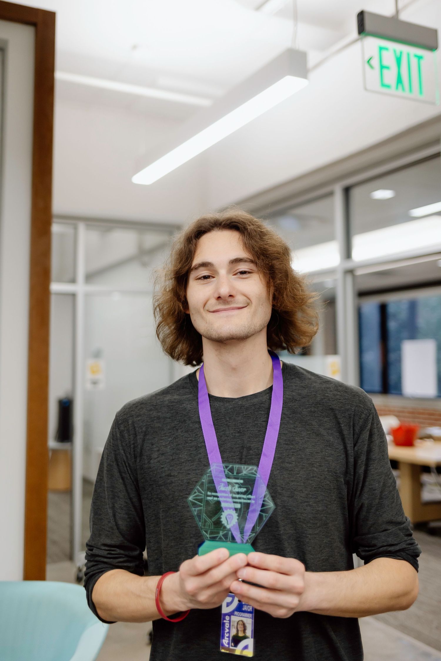 Someone wearing a dark gray shirt and purple lanyard smiles while holding something in an office setting near an exit sign.