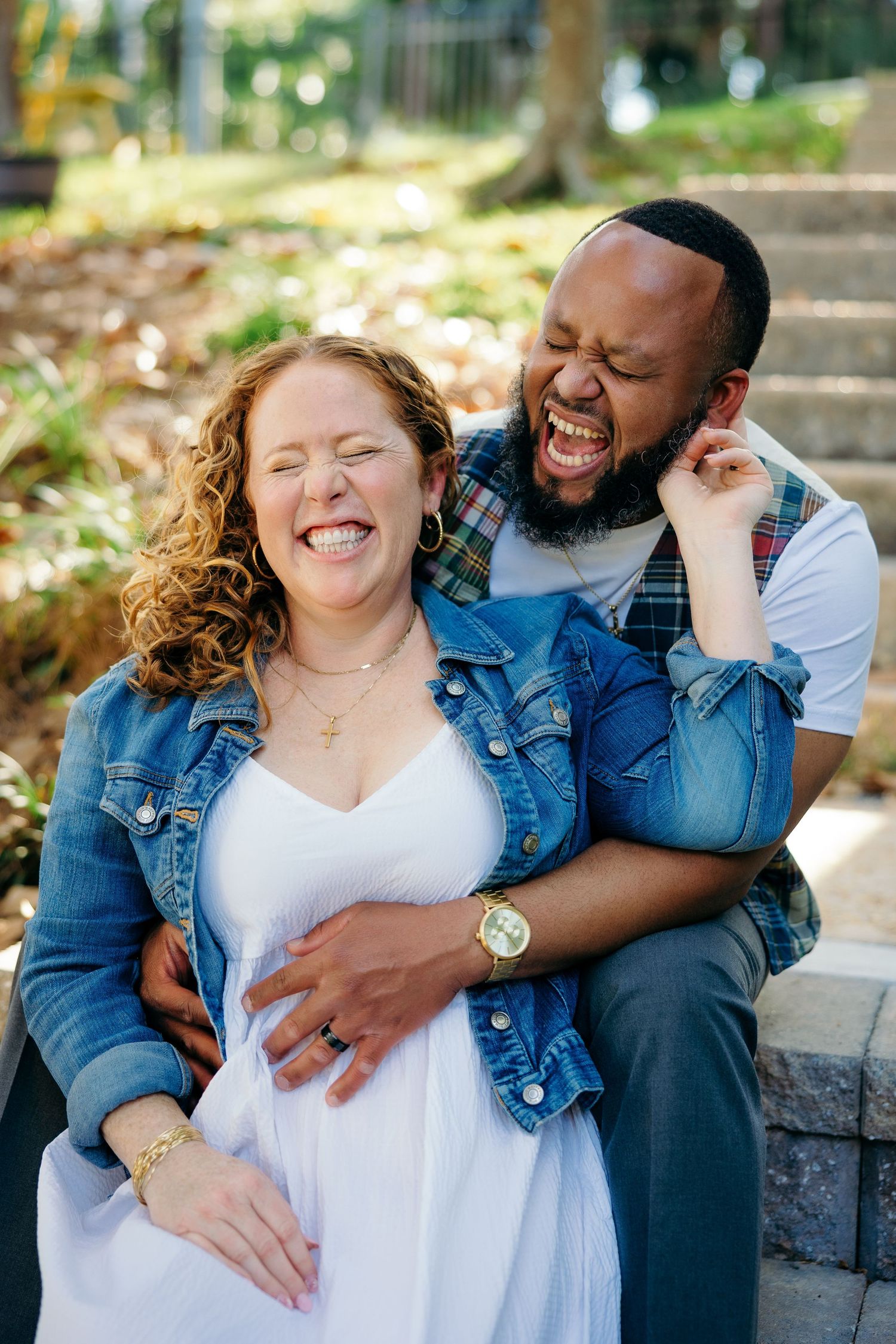 A joyful couple laughs together during an outdoor engagement photo session while wearing denim and white.