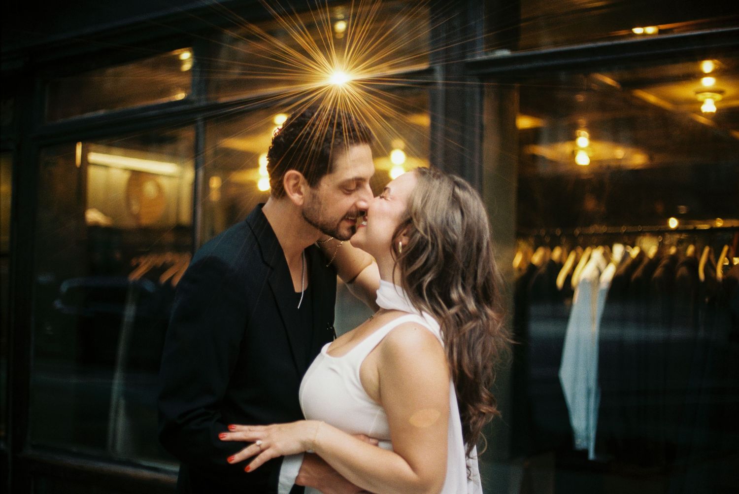 Romantic couple shares tender kisses in dimly lit evening setting with warm glowing lights in background.