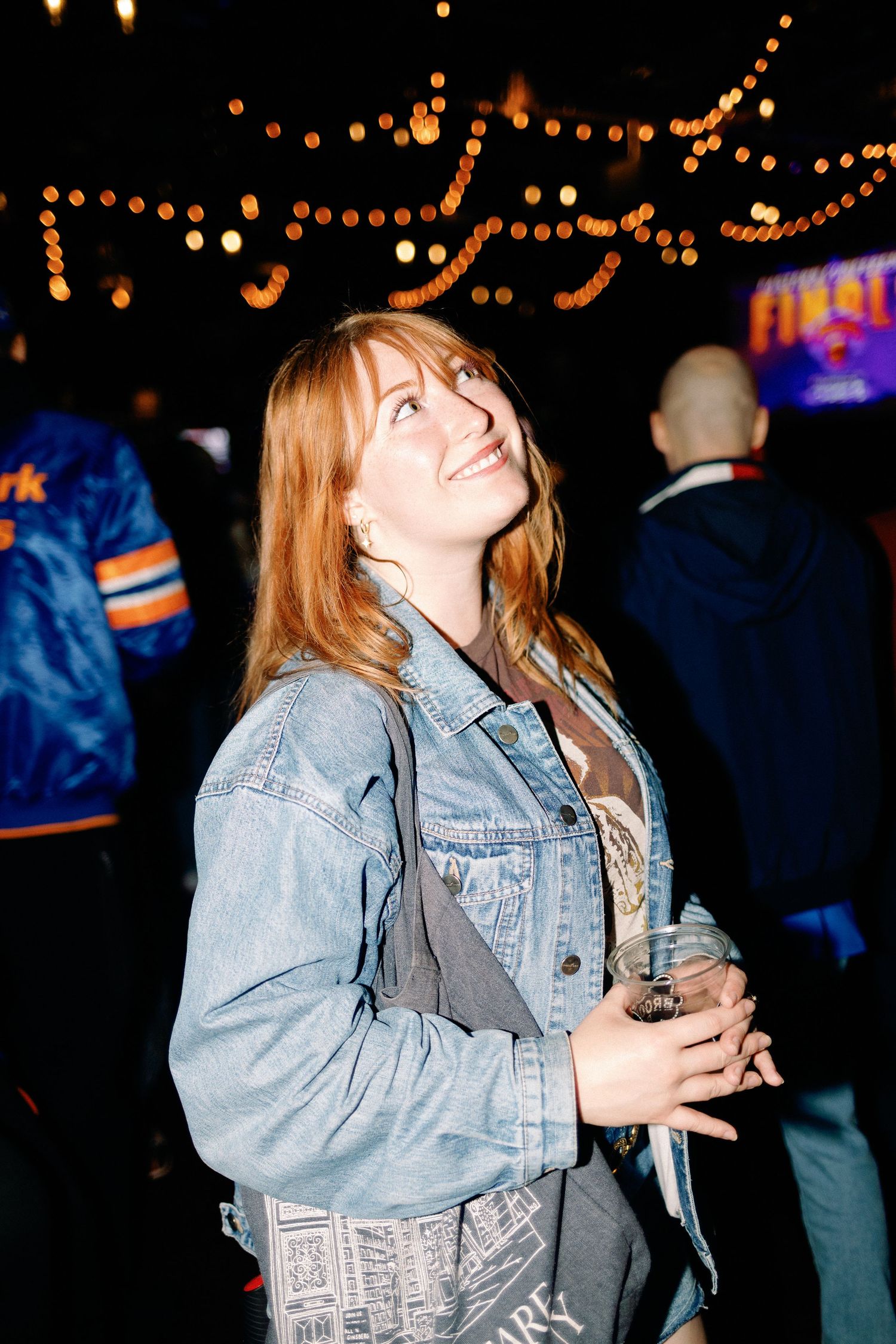 A smiling redhead woman in a denim jacket holds a drink at a festive night event with string lights.