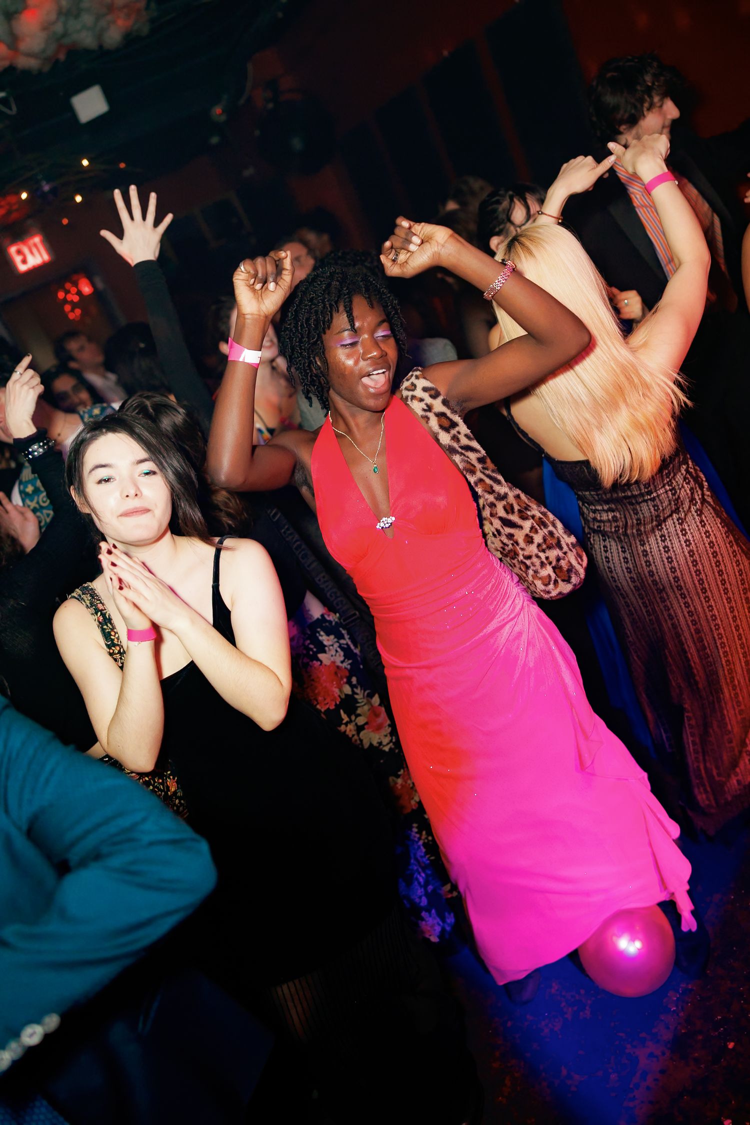 Young women dancing energetically at a party, wearing colorful dresses in a dimly lit nightclub venue.