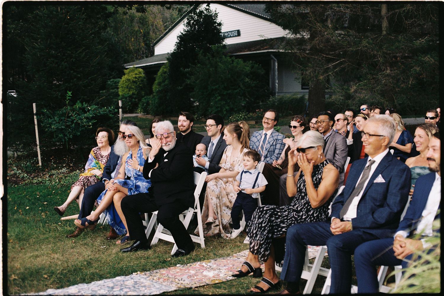 Wedding guests seated outdoors on white chairs during a garden ceremony surrounded by greenery and trees.