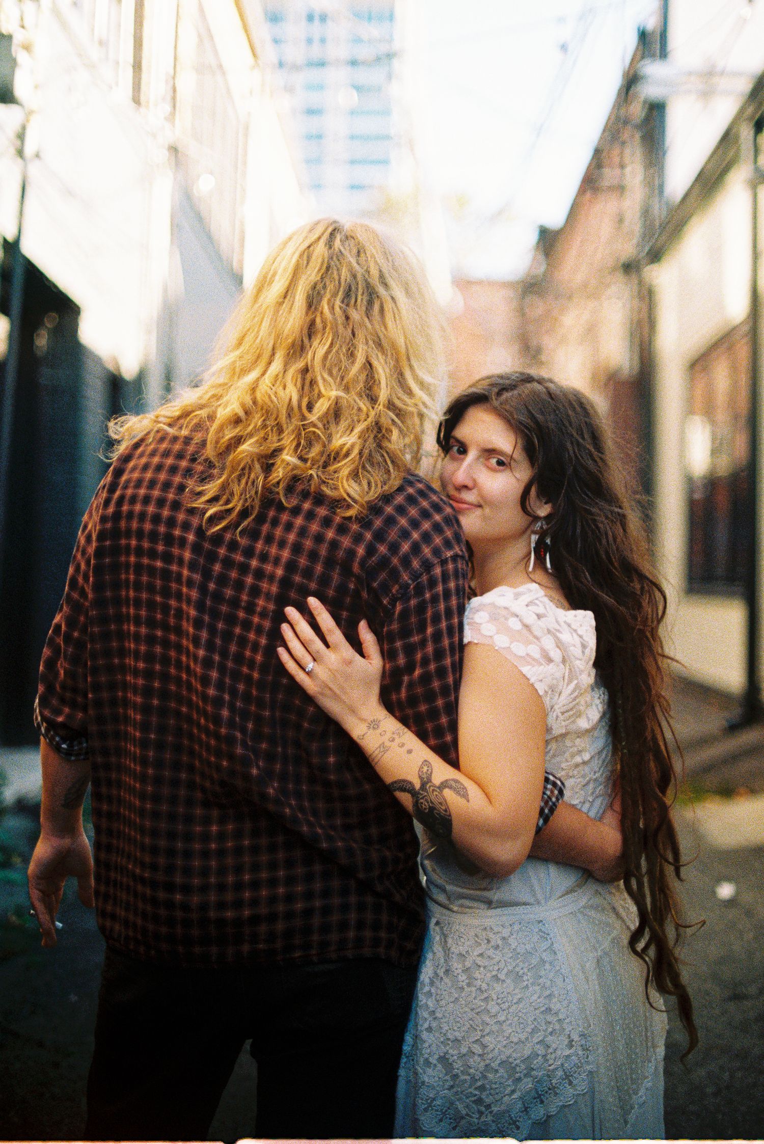 Couple embracing in a city alley, a blonde man in a plaid shirt facing away and a woman in a white lace dress with tattoos looking back.