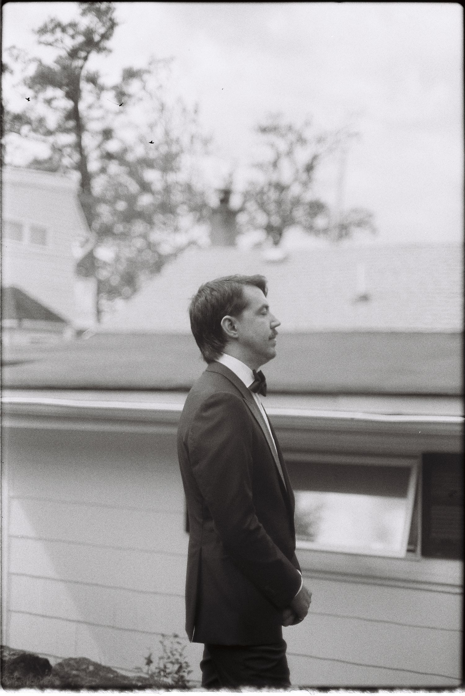 Black and white photo of a man in a tuxedo standing outdoors in profile view with houses and trees behind him.
