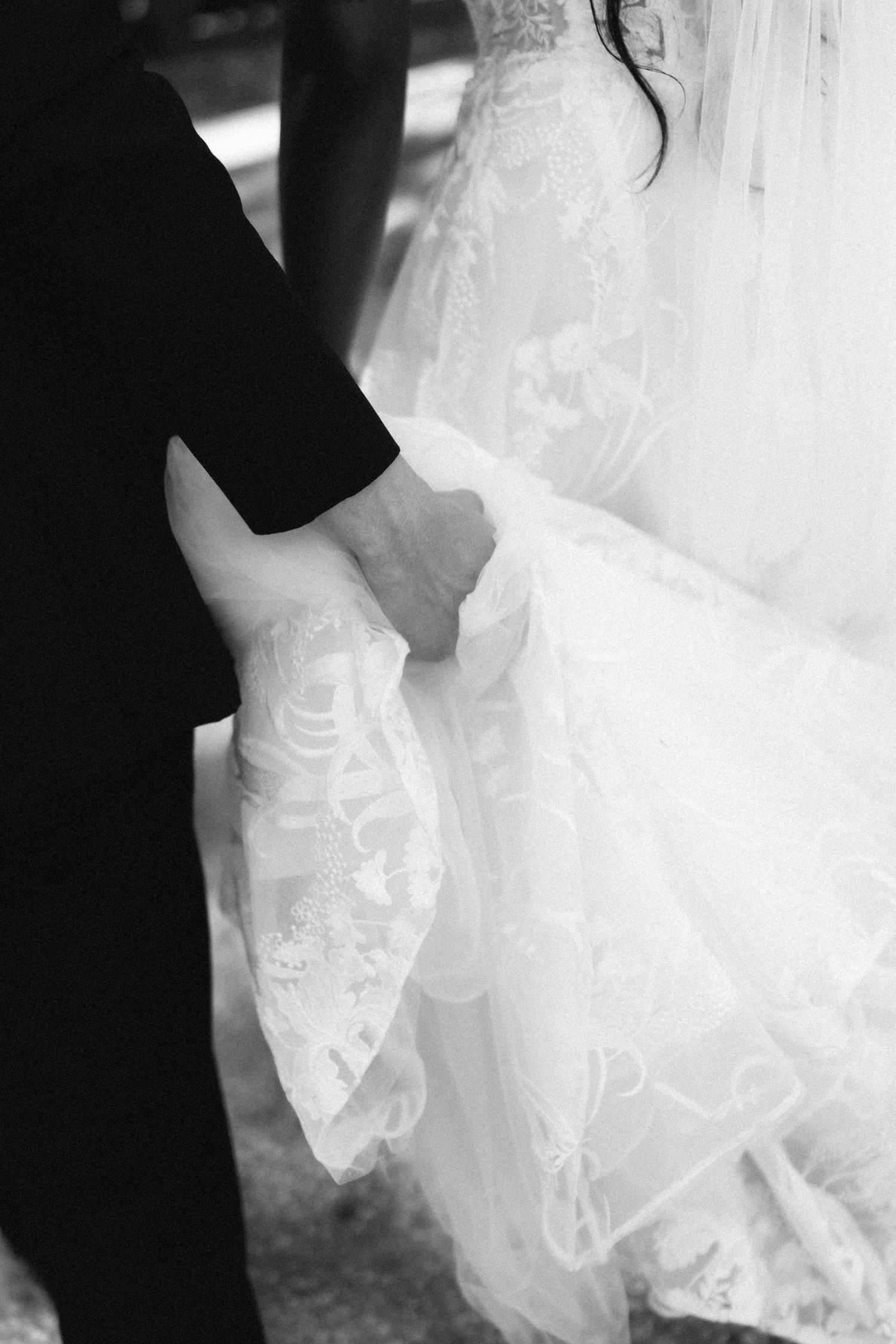 Black and white close-up of groom holding bride's hand beside her lace wedding gown.