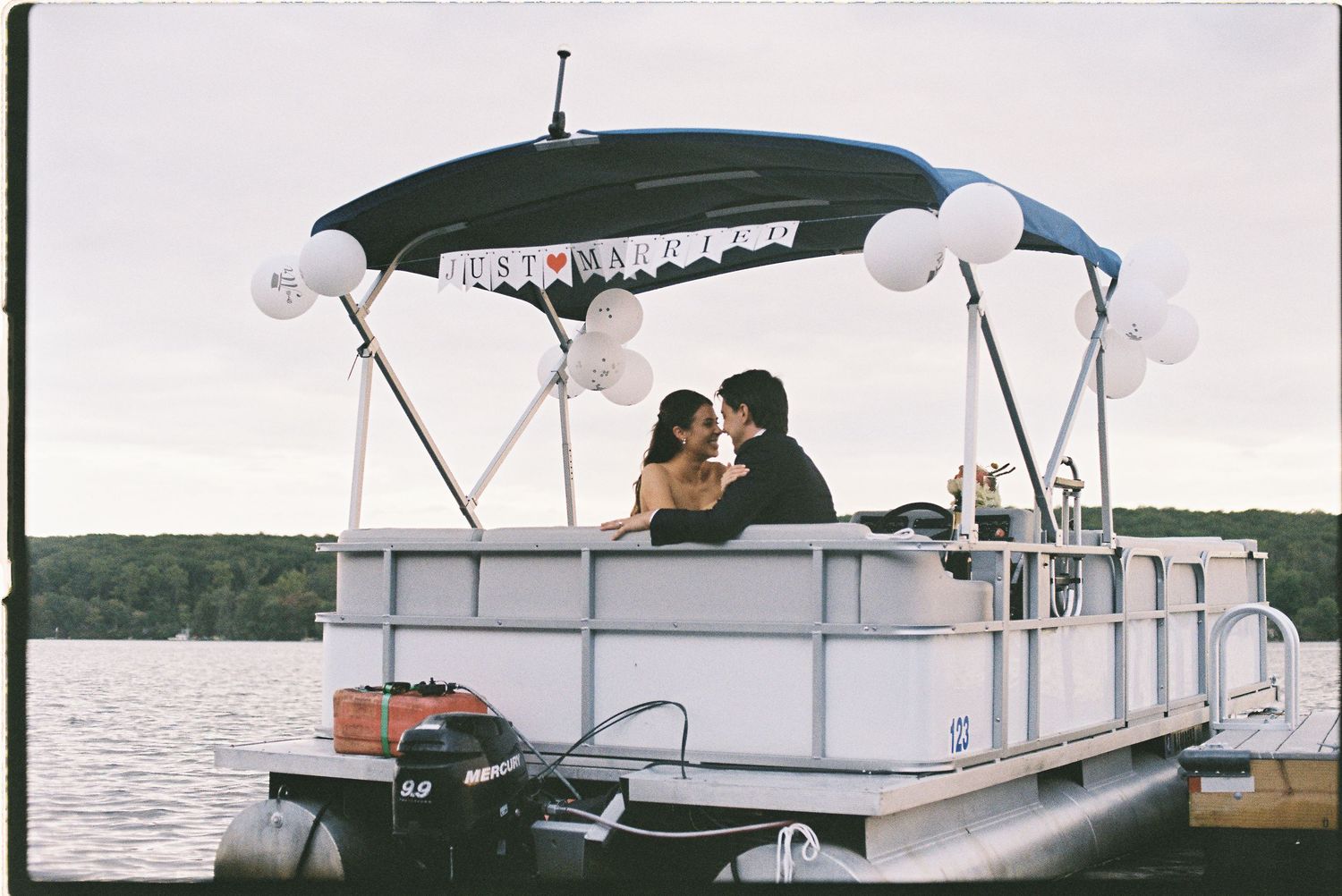 Couple kissing on a decorated pontoon boat with white balloons and bunting on a calm lake.
