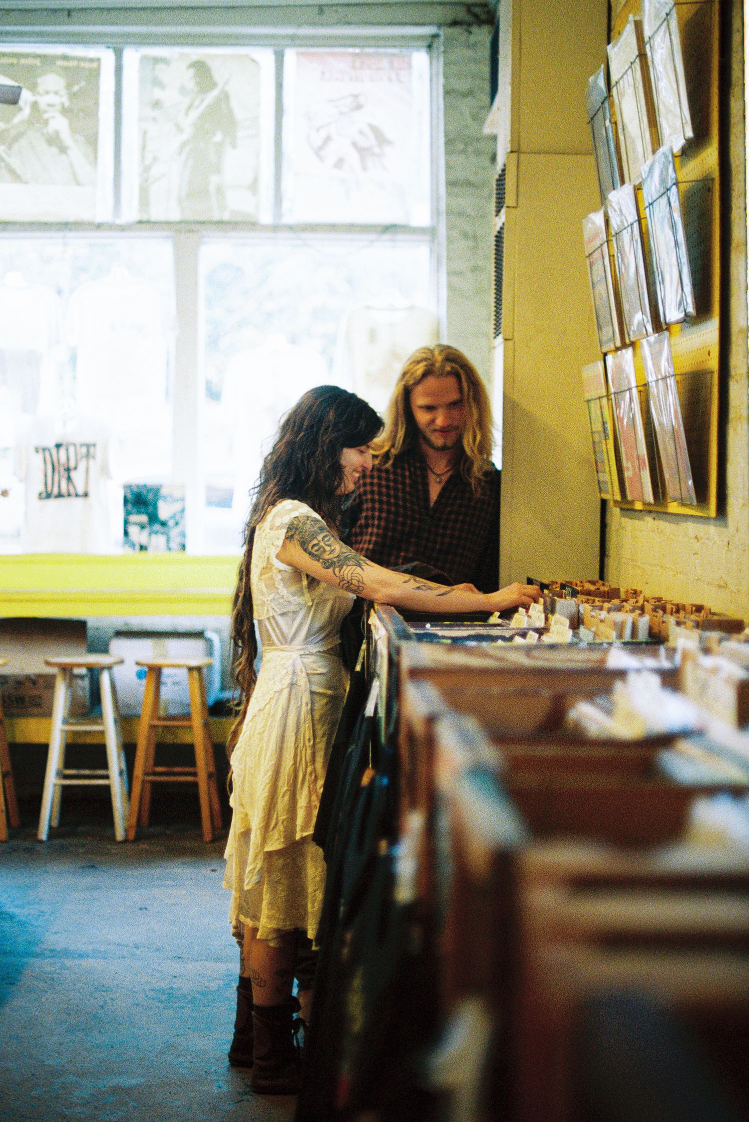 A couple browses vinyl records together in a sunlit record store with wooden stools and album covers displayed on the wall.