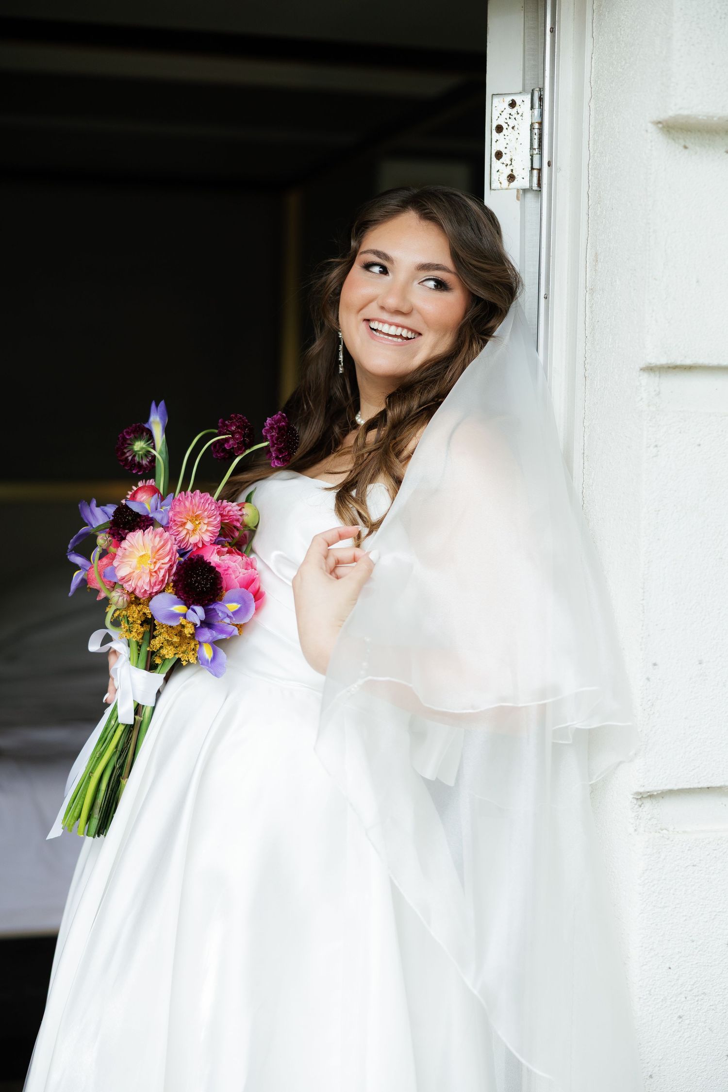 Smiling bride in white gown and veil holds colorful bouquet of pink, purple, and blue flowers while leaning against a white door.