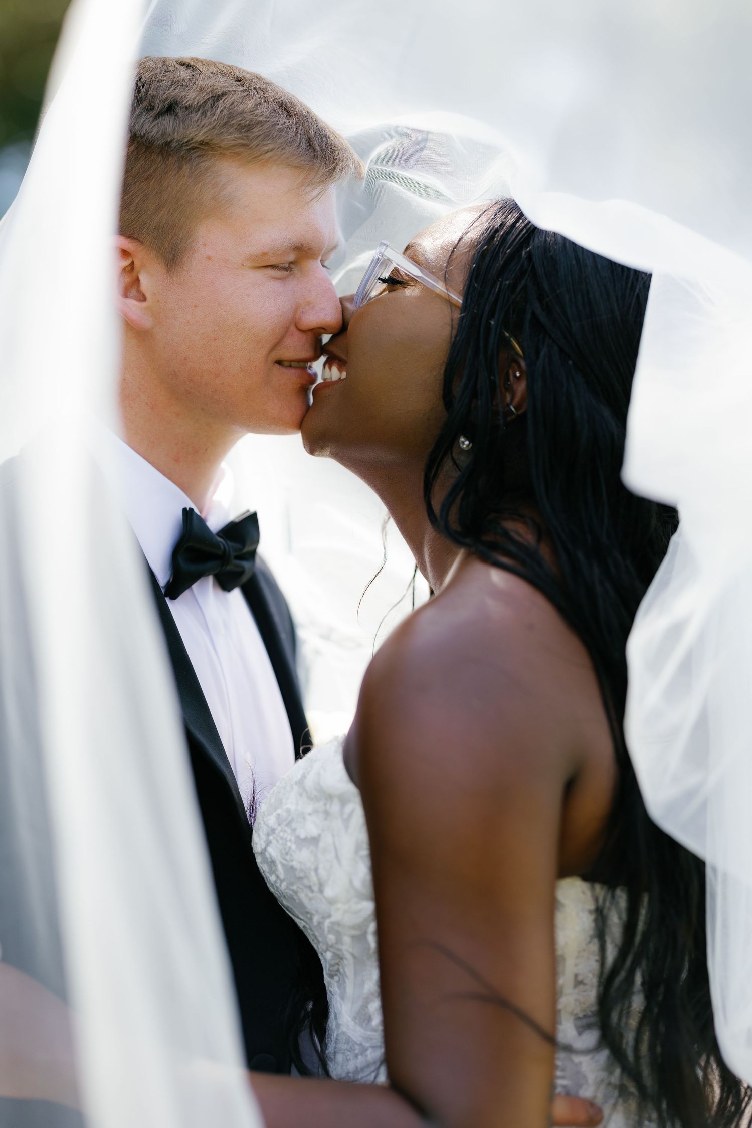 Interracial bride and groom sharing a romantic kiss under a flowing white veil on their wedding day.