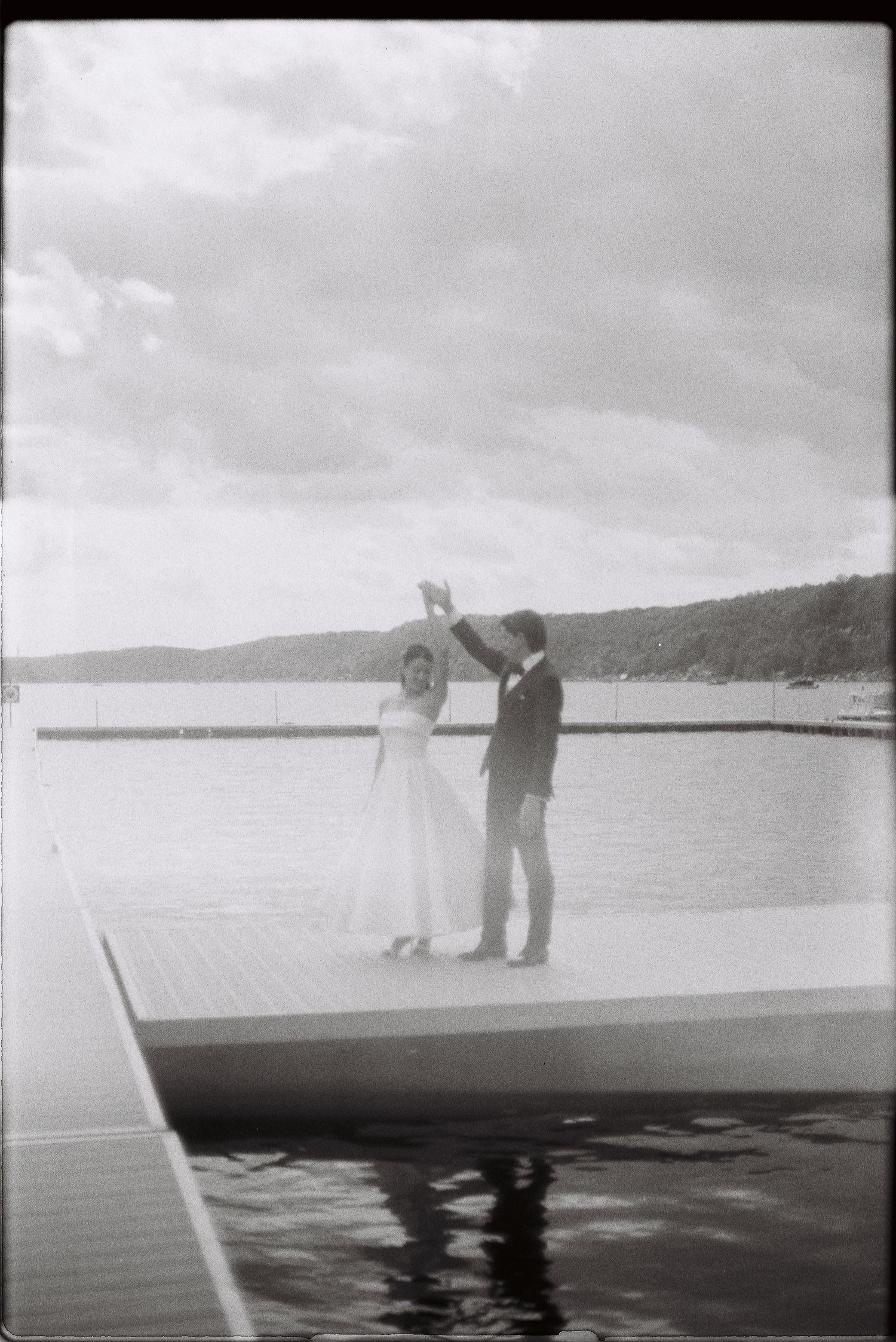 Black and white photo of bride and groom dancing on lakeside dock.