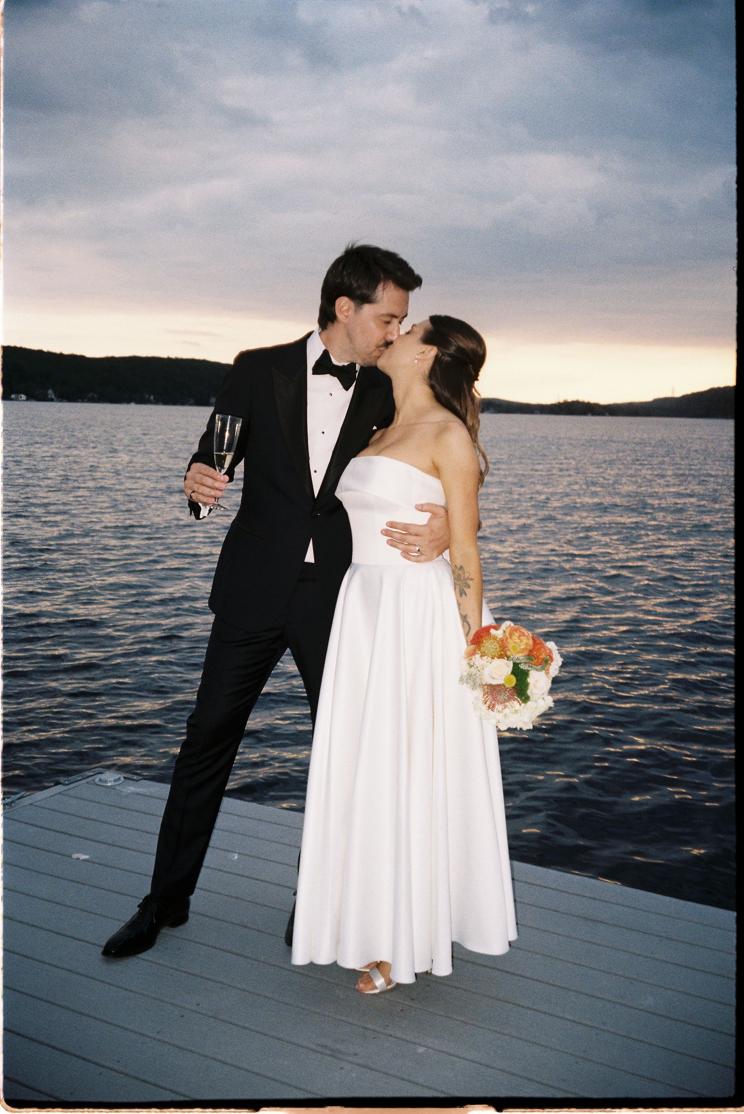 Newlyweds kiss on dock at sunset, groom holds champagne, bride holds bouquet.