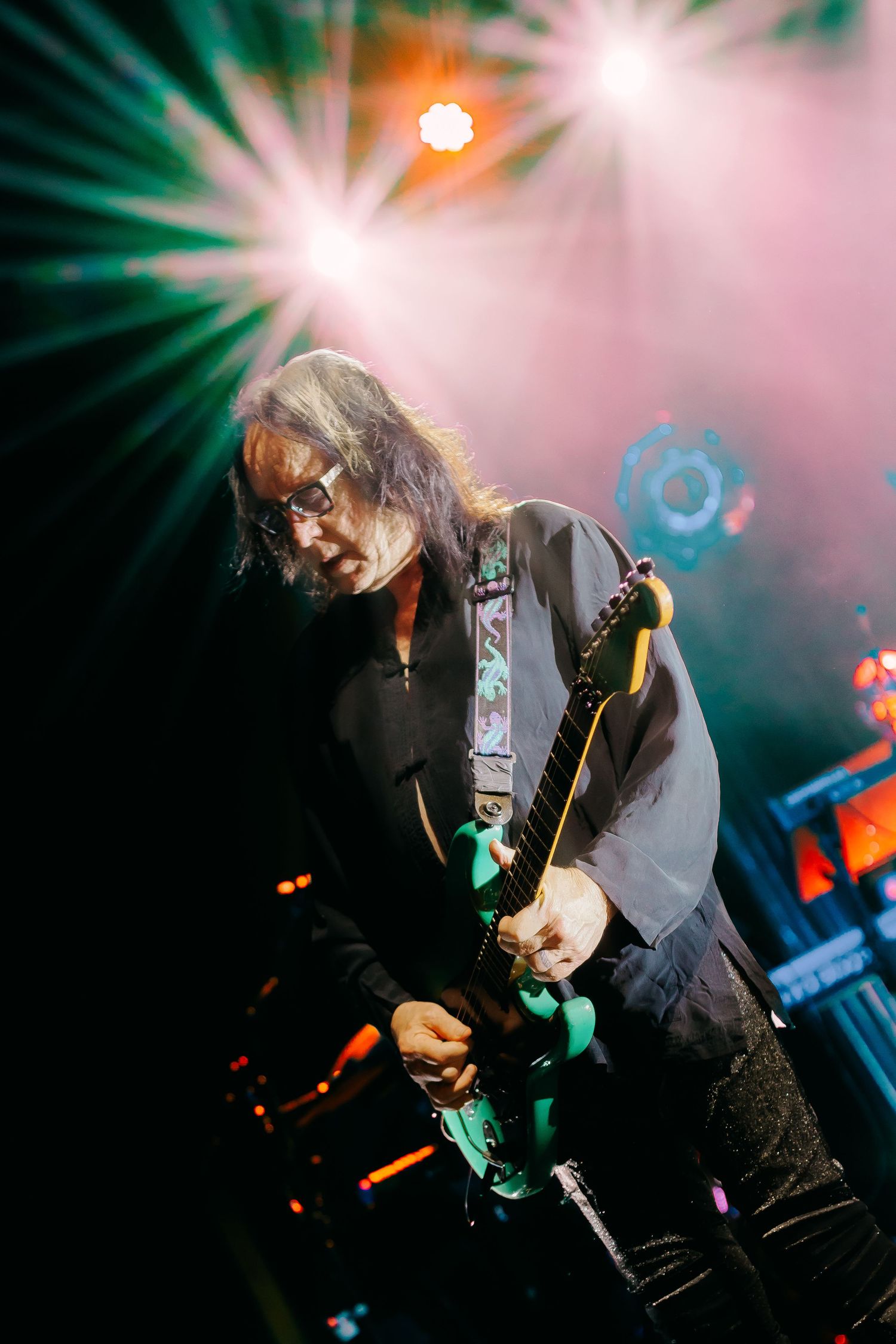 Dynamic concert photograph of a musician playing guitar on stage with dramatic lighting and smoke effects in the background.