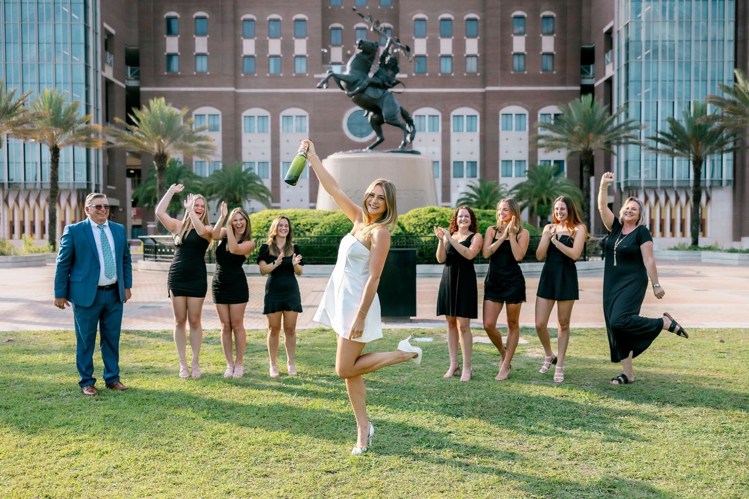 A group of people in formal attire pose together in a progression of jumping photos on a grassy lawn.